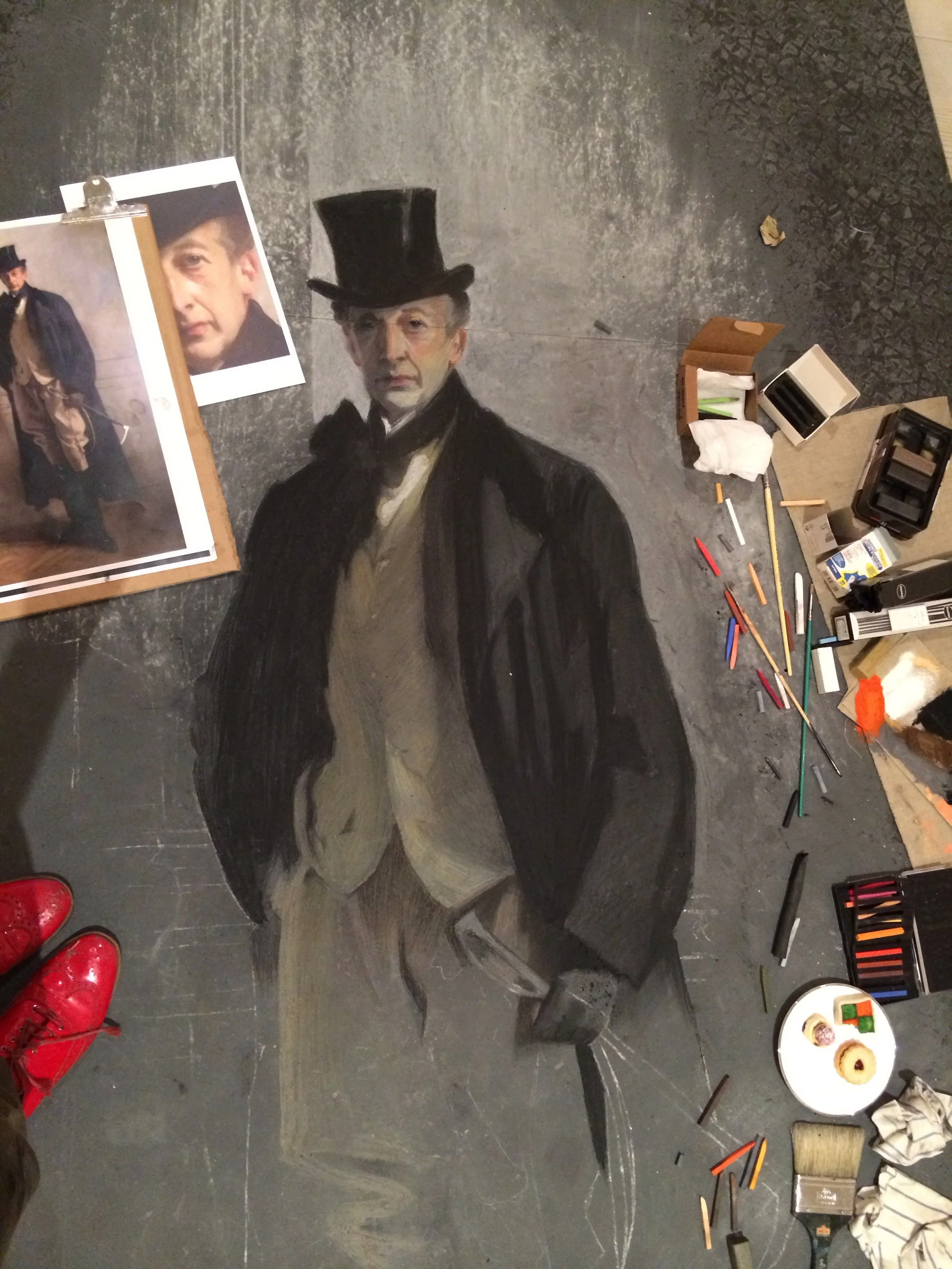 A partially completed chalk art of a man in period clothing, with an additional photo of the same man on a clipboard nearby, and art supplies scattered around on the floor.