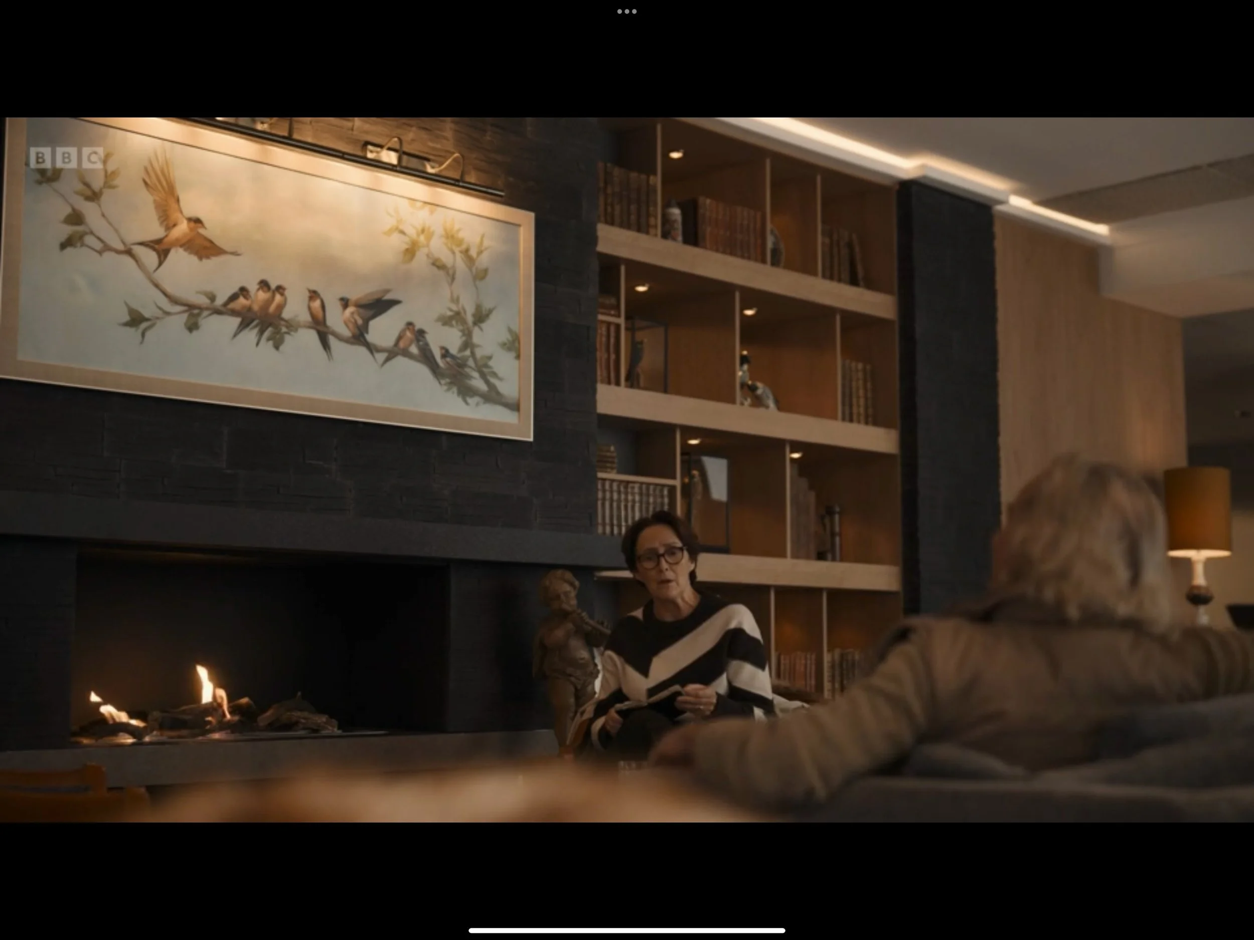Two women having a conversation in a modern living room with a fireplace, bookshelf, and large painting of birds on a branch