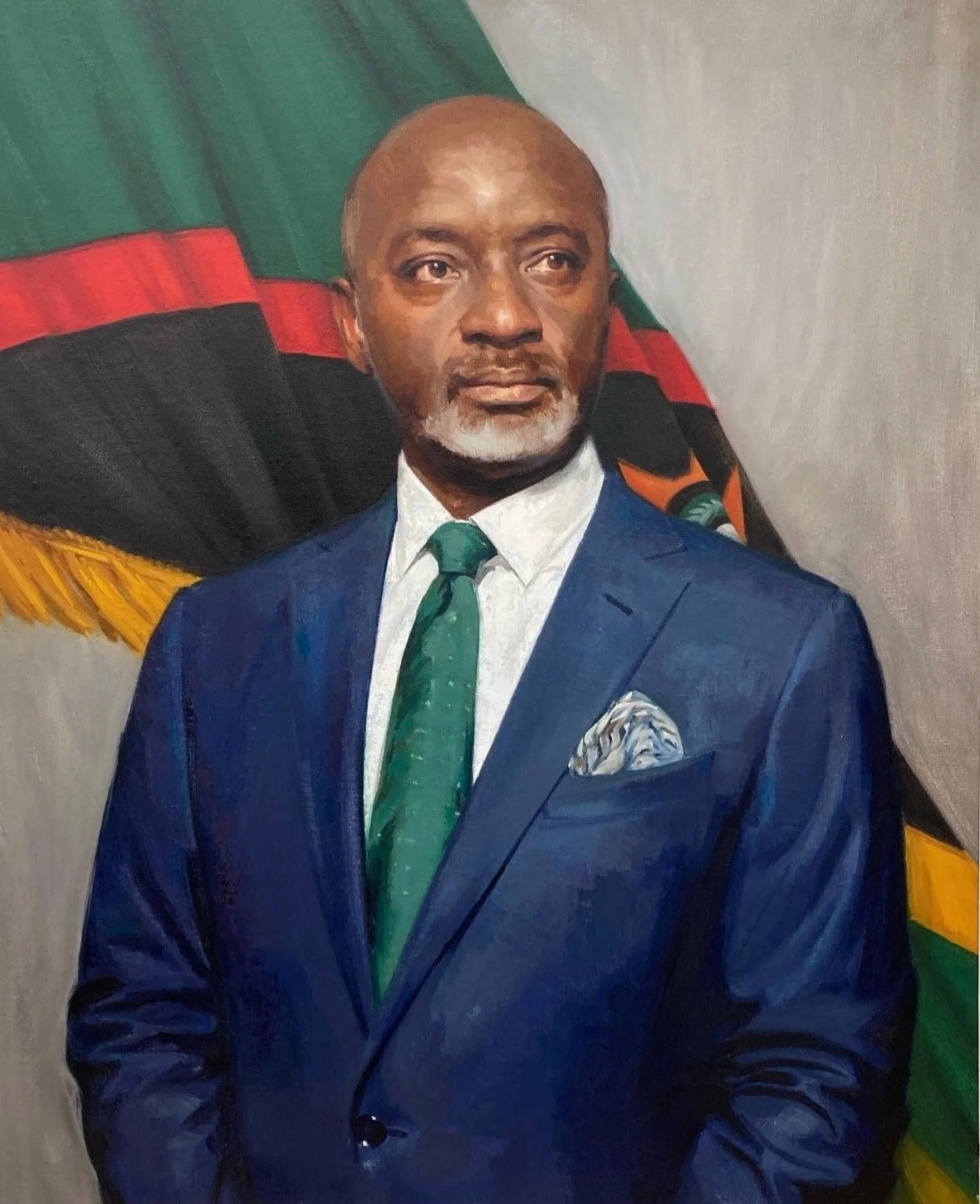 Portrait of a man in a blue suit, white shirt, and green tie, standing in front of a flag with green, black, red, and yellow colors.