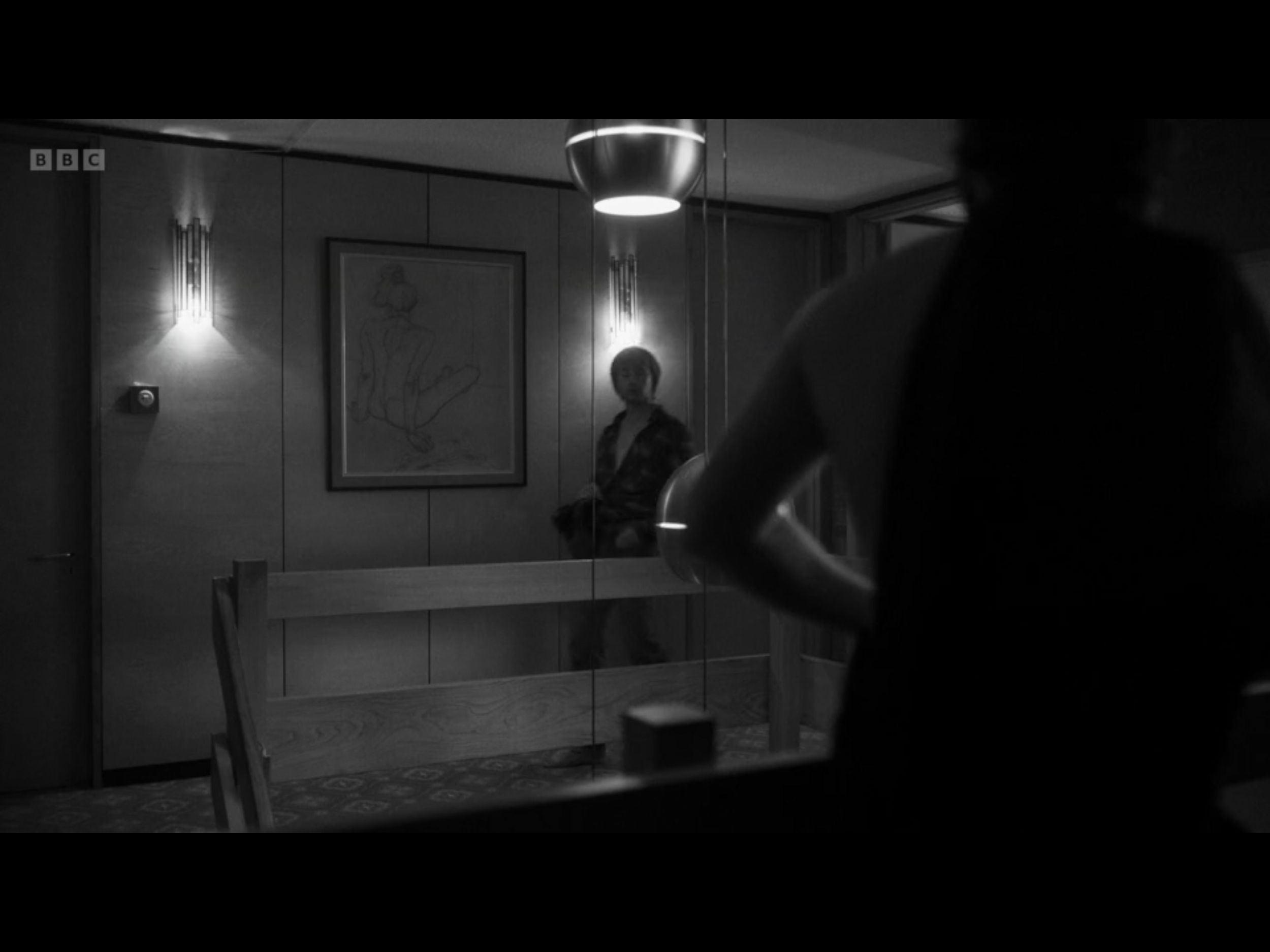 A black and white photo of a woman sitting on a ledge in a room with wooden walls and a framed drawing of a woman on the wall. A person is partially visible in the foreground, with their arm and shoulder in focus.