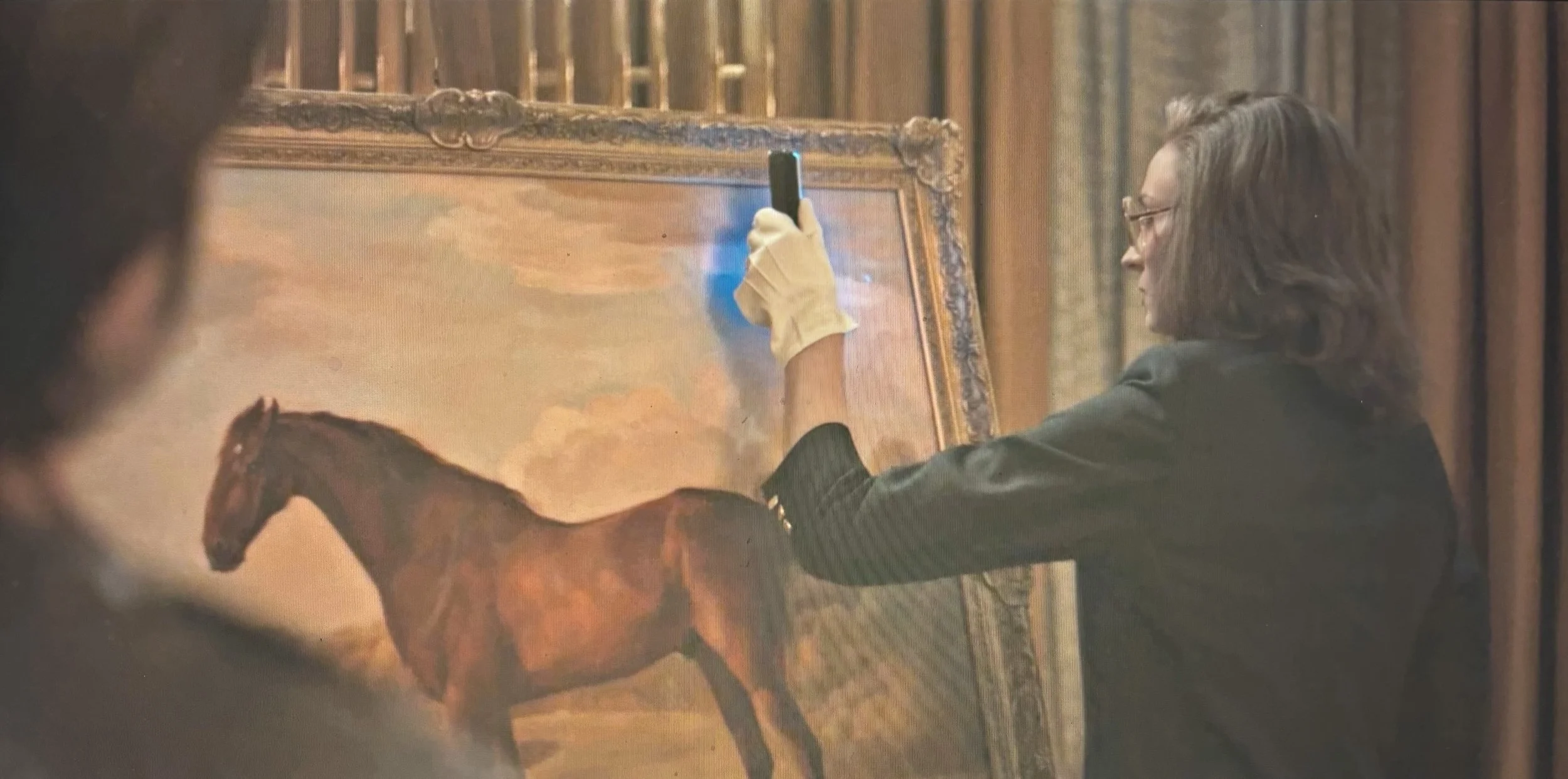 A woman with glasses and dark hair cleaning a framed painting of a brown horse in an ornate gold frame, while wearing yellow gloves.