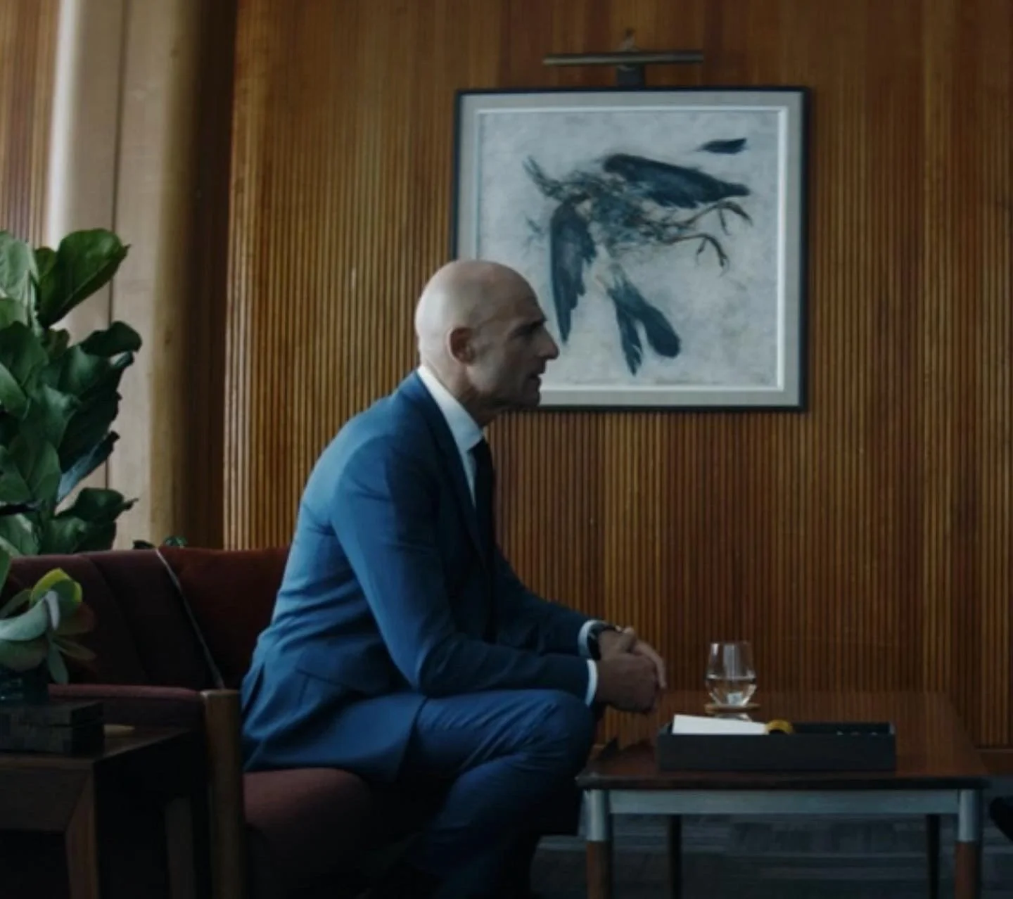 A bald man in a blue suit sitting in a room with a wooden wall, a painting of abstract birds behind him, a green plant, a glass of water, and a black box on a small table.