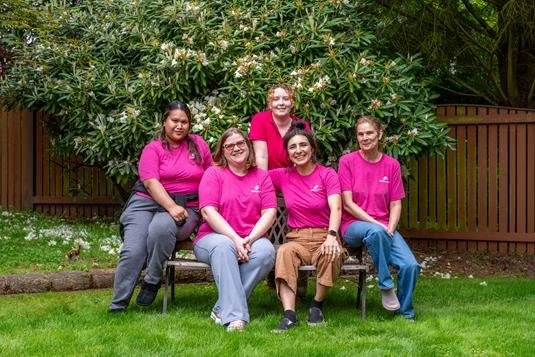 A group of five women sitting and standing on a bench in a garden, surrounded by green trees and a wooden fence, all wearing matching pink shirts.