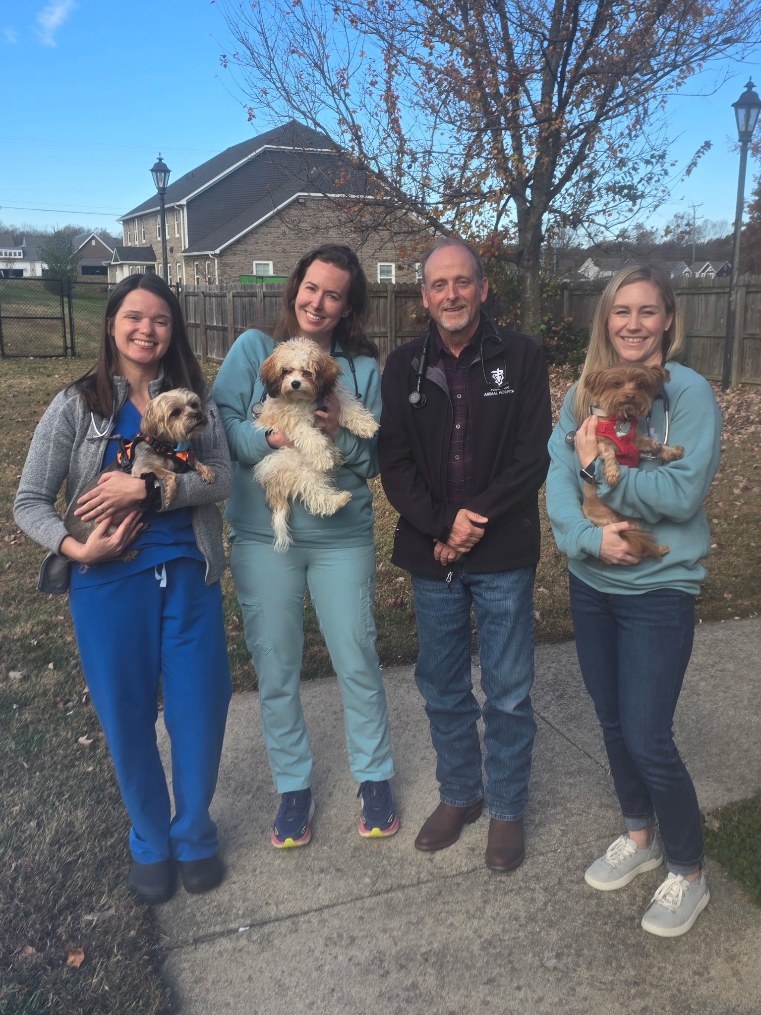 Four people, three women and one man, standing outdoors on a sidewalk in front of a wooden fence and a house with a tree behind them. The women are holding small dogs, and the man is standing between two women. The group is smiling and dressed casually.