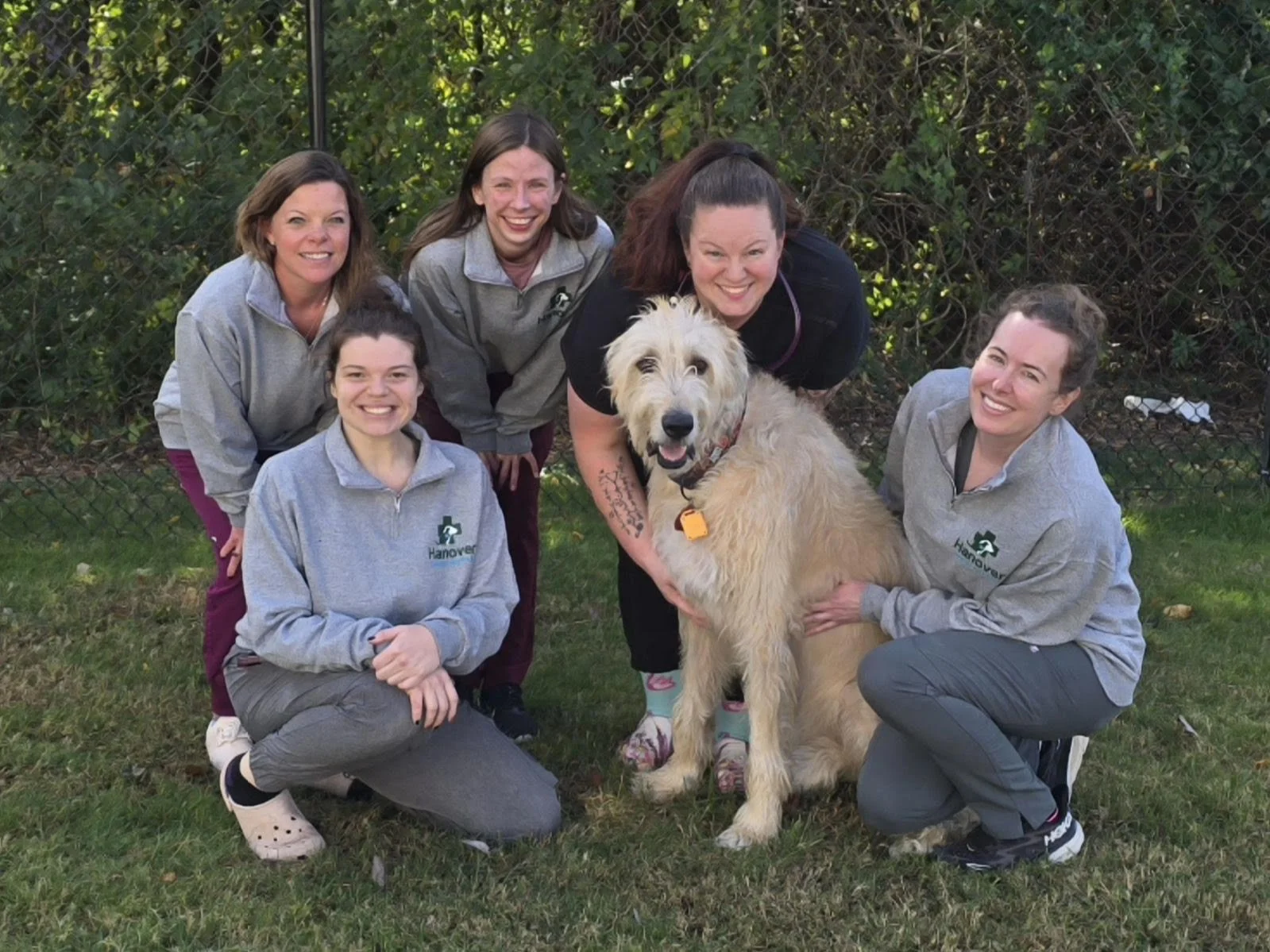 Our Staff: Five women and one large dog with light-colored fur outdoors in front of a chain-link fence, all smiling at the camera.