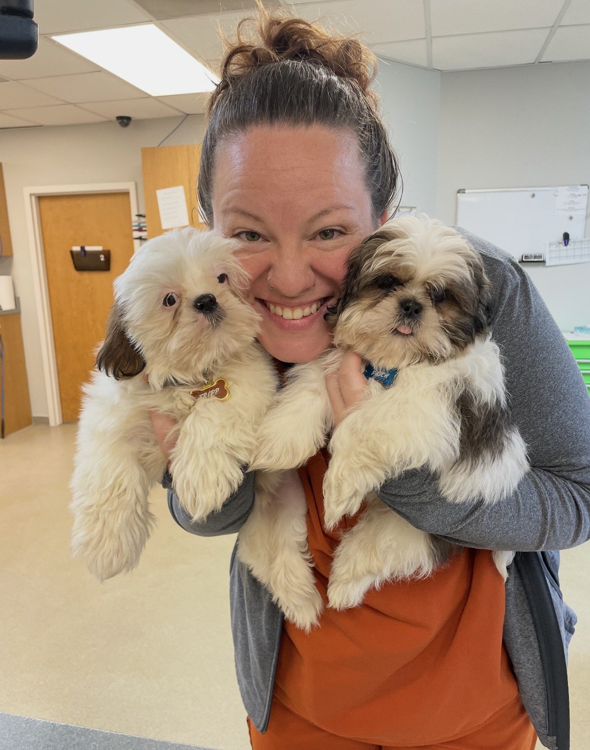 A woman smiling and holding two adorable fluffy puppies indoors, likely in an office or clinic setting.