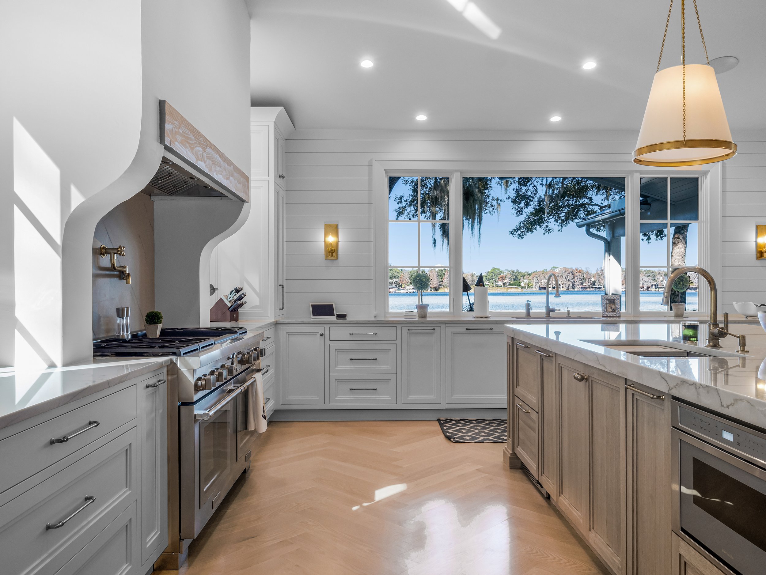 Bright kitchen with white cabinetry, marble countertops, and a large window overlooking a lake with trees. Modern appliances, gold fixtures, and a hanging pendant light are visible.