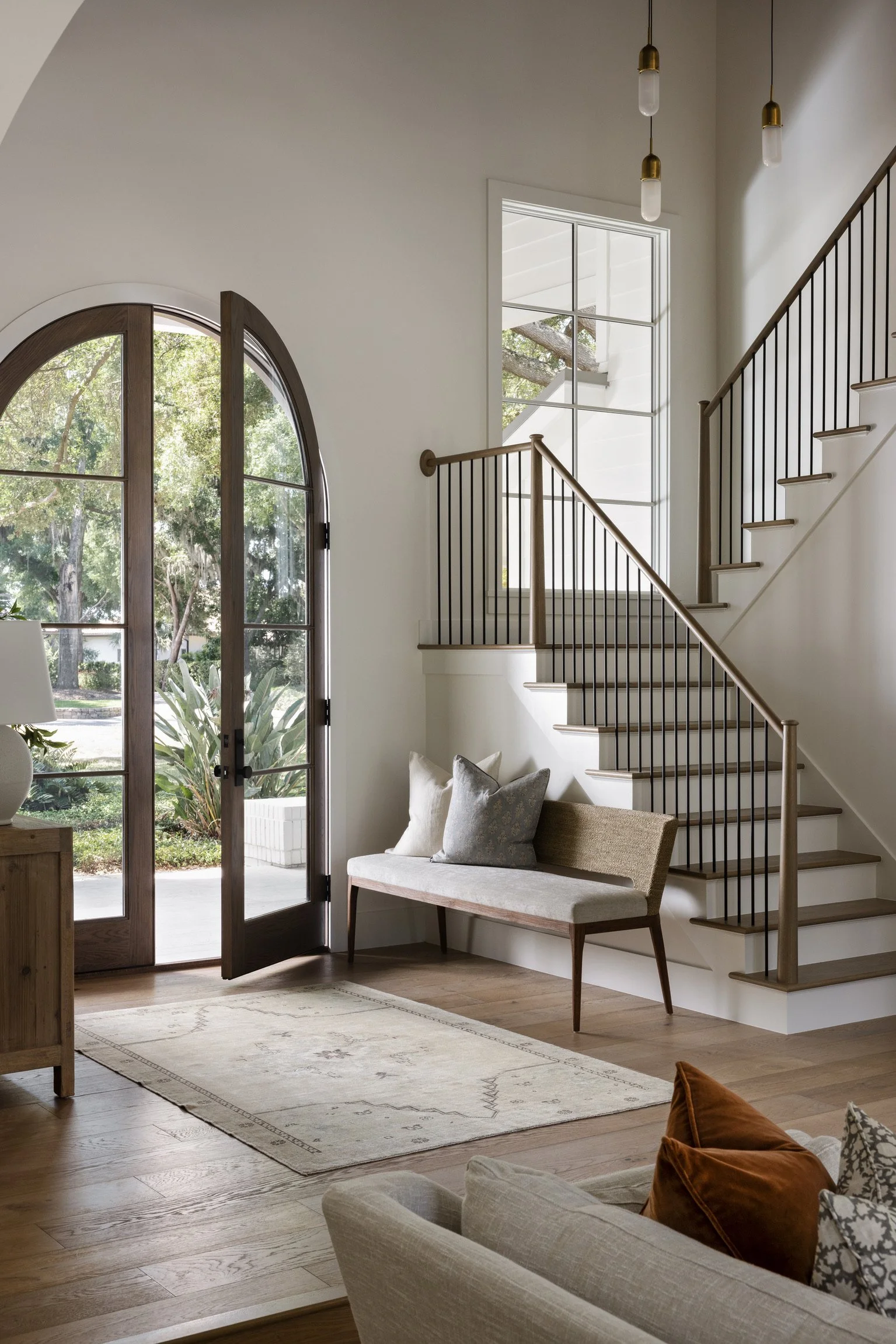 Interior of a modern living room with wooden floors, a bench with cushions, a large arched glass door, and a staircase with a black railing.