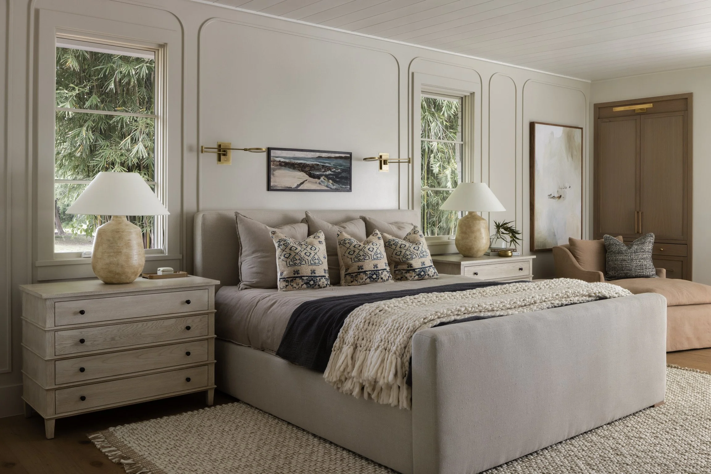 A neatly made bedroom with a beige upholstered bed, decorative pillows, matching nightstands with lamps, two windows showing greenery outside, and a sitting area with an armchair and artwork on the wall.