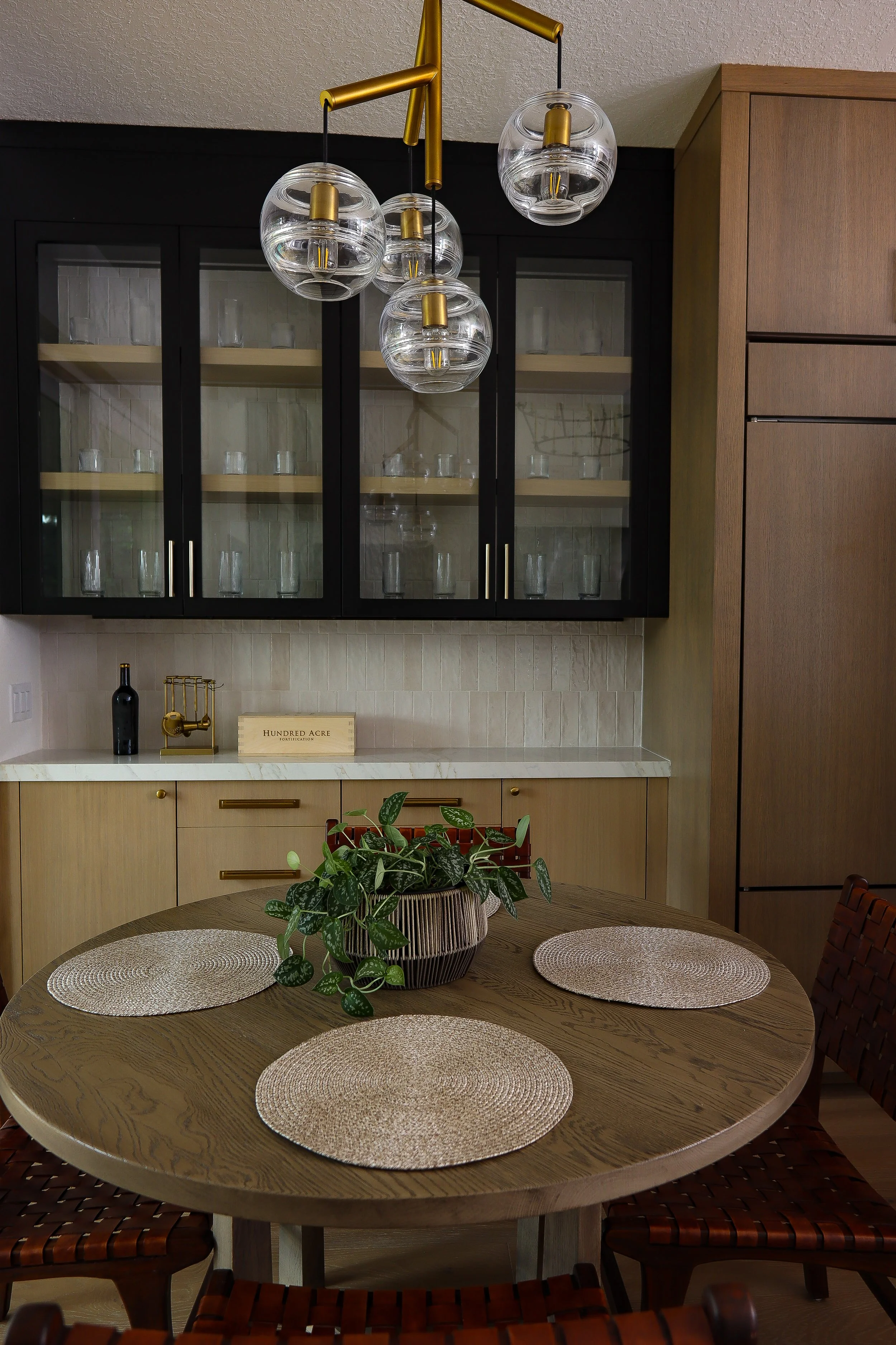 A dining room with a wooden oval table decorated with a potted plant and three circular woven placemats. A black and gold glass chandelier hangs above, and in the background, there are black glass-front kitchen cabinets with glasses inside, beige cabinetry with gold handles, and decorative items on the counter.