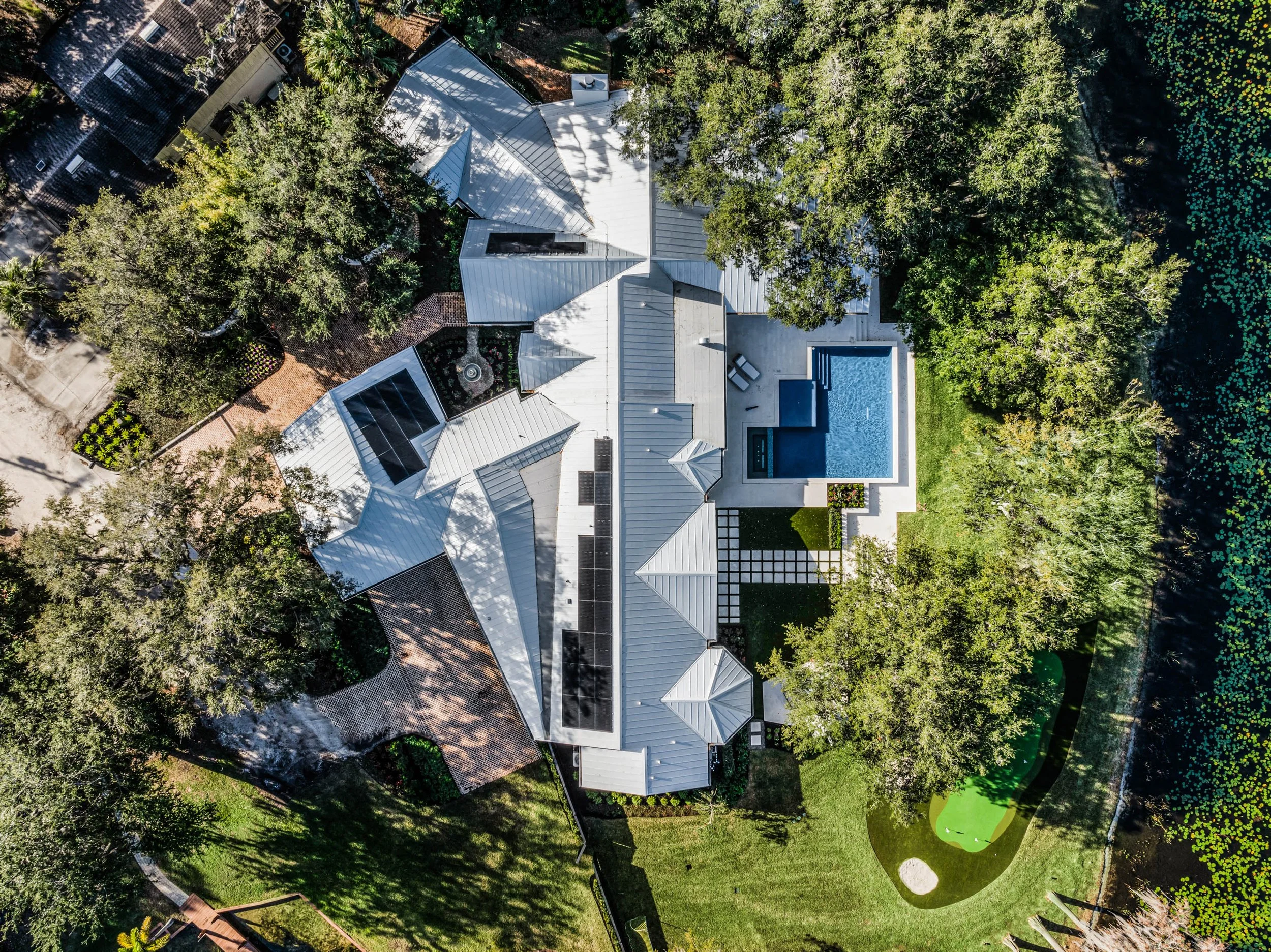 Aerial view of a modern house with white metal roof, surrounded by lush green trees, a swimming pool, a patio with furniture, a putting green, and landscaped yard.