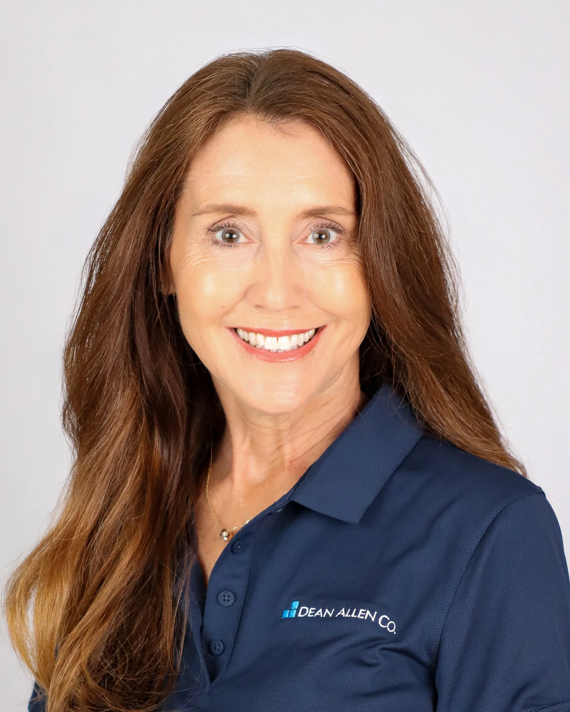 A smiling woman with long auburn hair wearing a navy blue polo shirt with 'Dean Allen Co.' embroidered on it, posing against a plain light background.