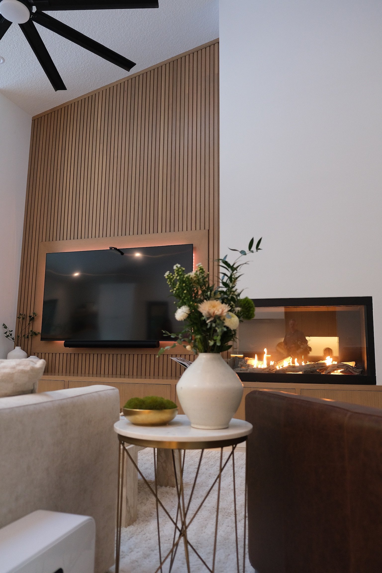 Living room with a flat-screen TV on a wooden accent wall, a fireplace, a white vase with flowers on a small round table, and a beige sofa.