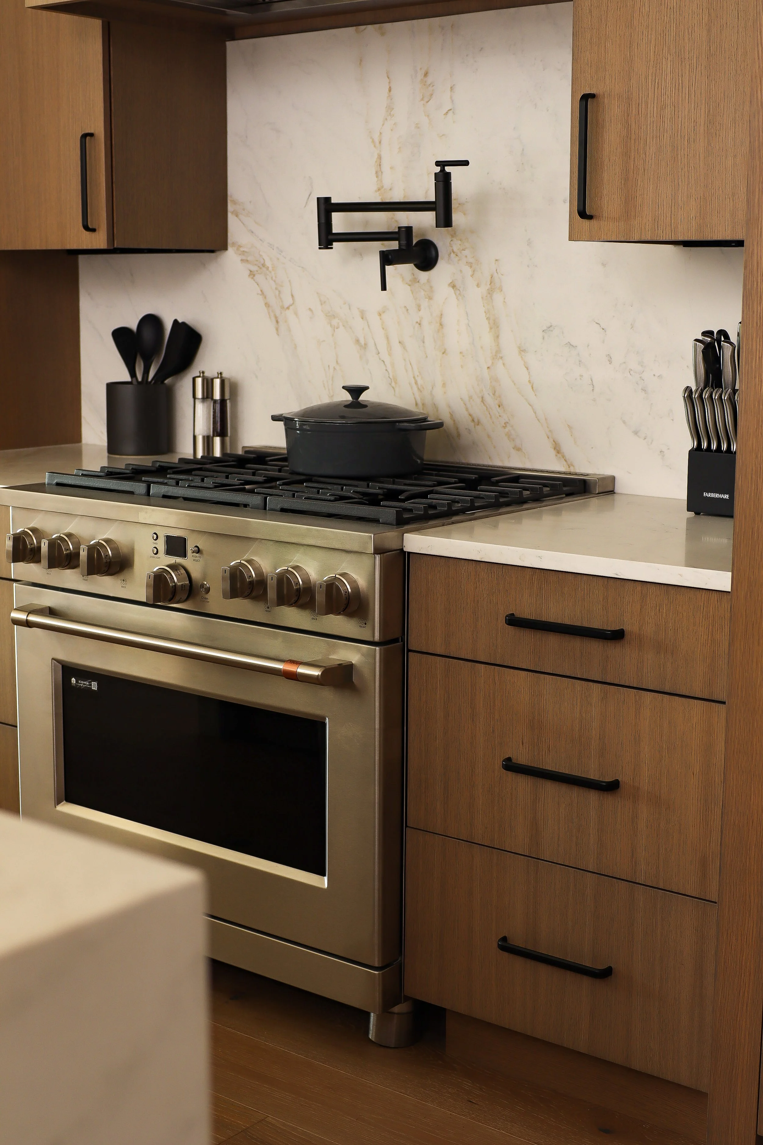 A modern kitchen with a stainless steel oven, a black pot on the stove, a black faucet, a utensil holder with black utensils, and a knife block on the counter.