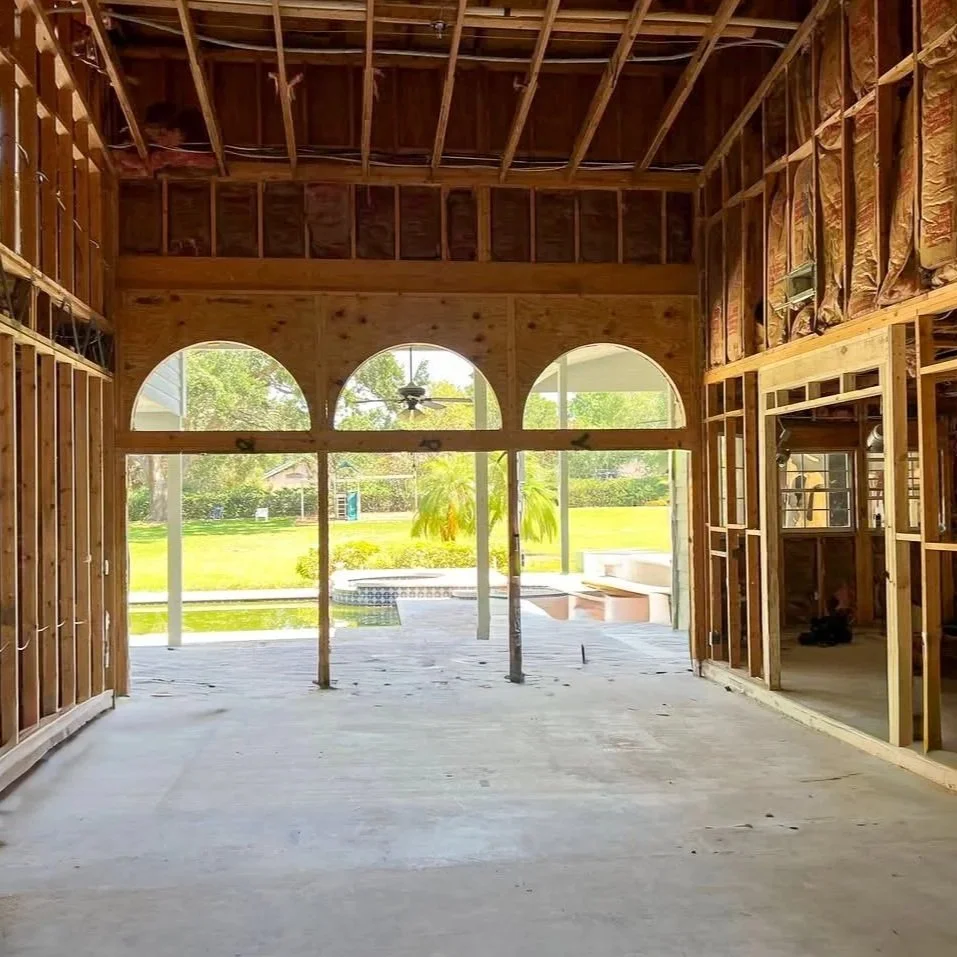 Interior of a house under construction, showing exposed wooden framing, large arched windows, and a view of a backyard with a pool and green lawn.