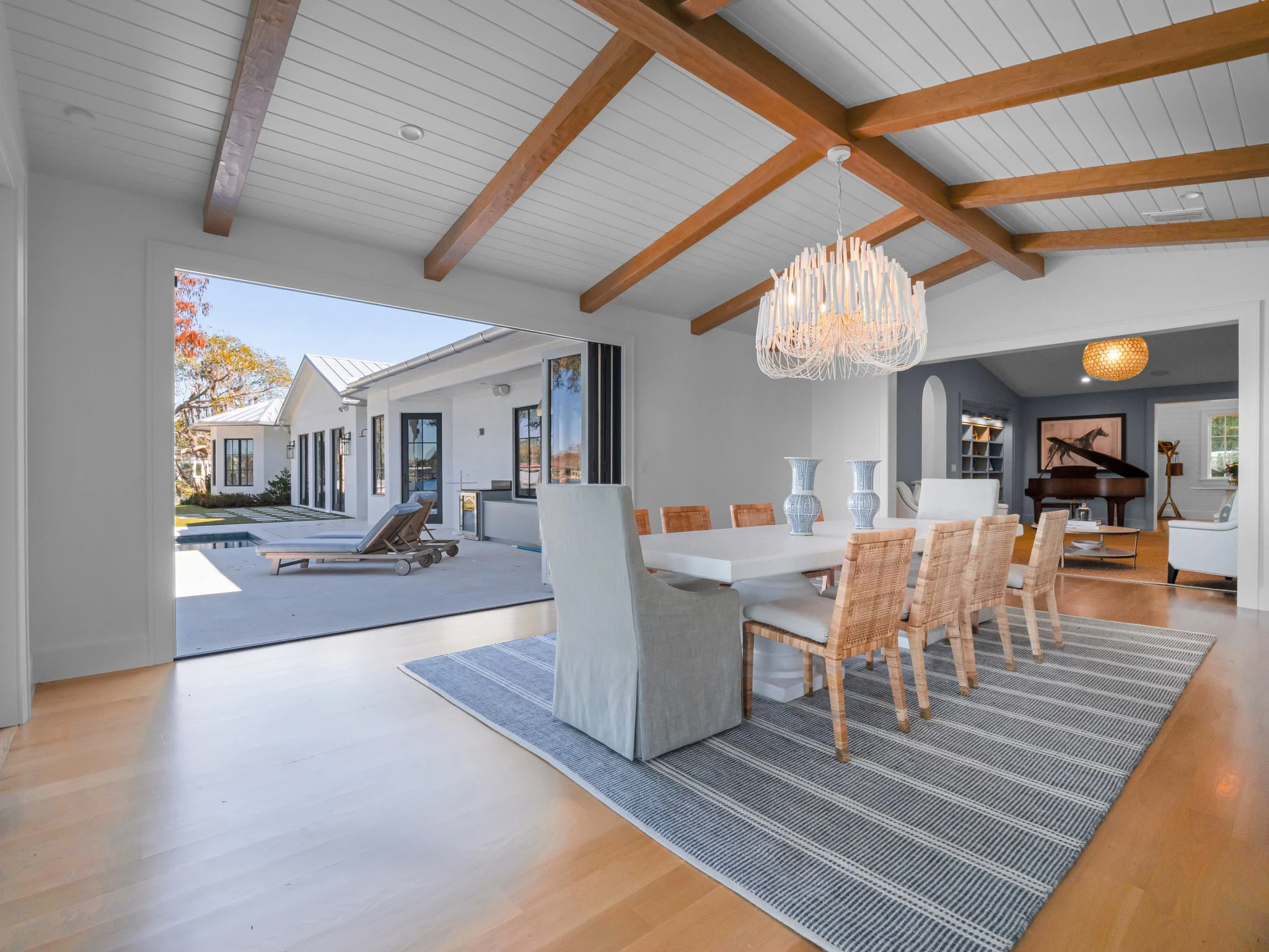Open-concept dining room with wooden beams on white ceiling, large white dining table with eight chairs, three blue vases on the table, and a modern chandelier. Sliding glass door opens to outdoor patio with lounge chairs, pool, and yard. Adjacent ro