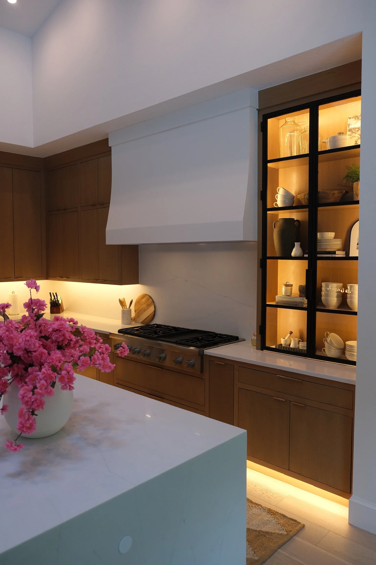 Modern kitchen with white countertops, wooden cabinets, and a kitchen range. A decorative display cabinet with glass doors and warm interior lighting holds dishes and vases. A white vase with pink flowers is on the island in the foreground.