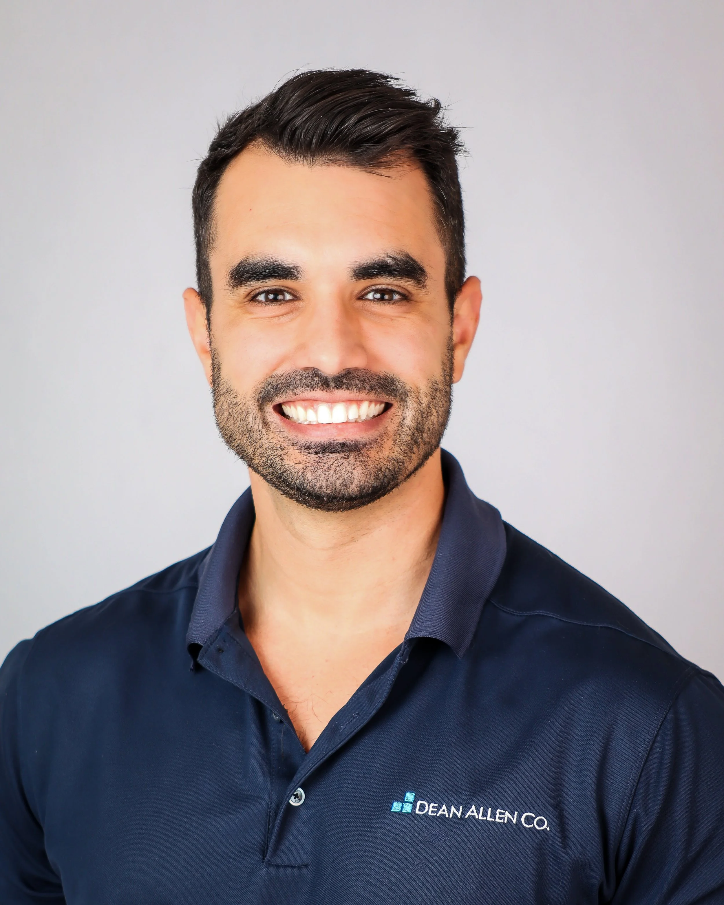 A smiling man with dark hair, beard, wearing a navy blue polo shirt with 'Dean Allen Co.' logo, against a plain light background.