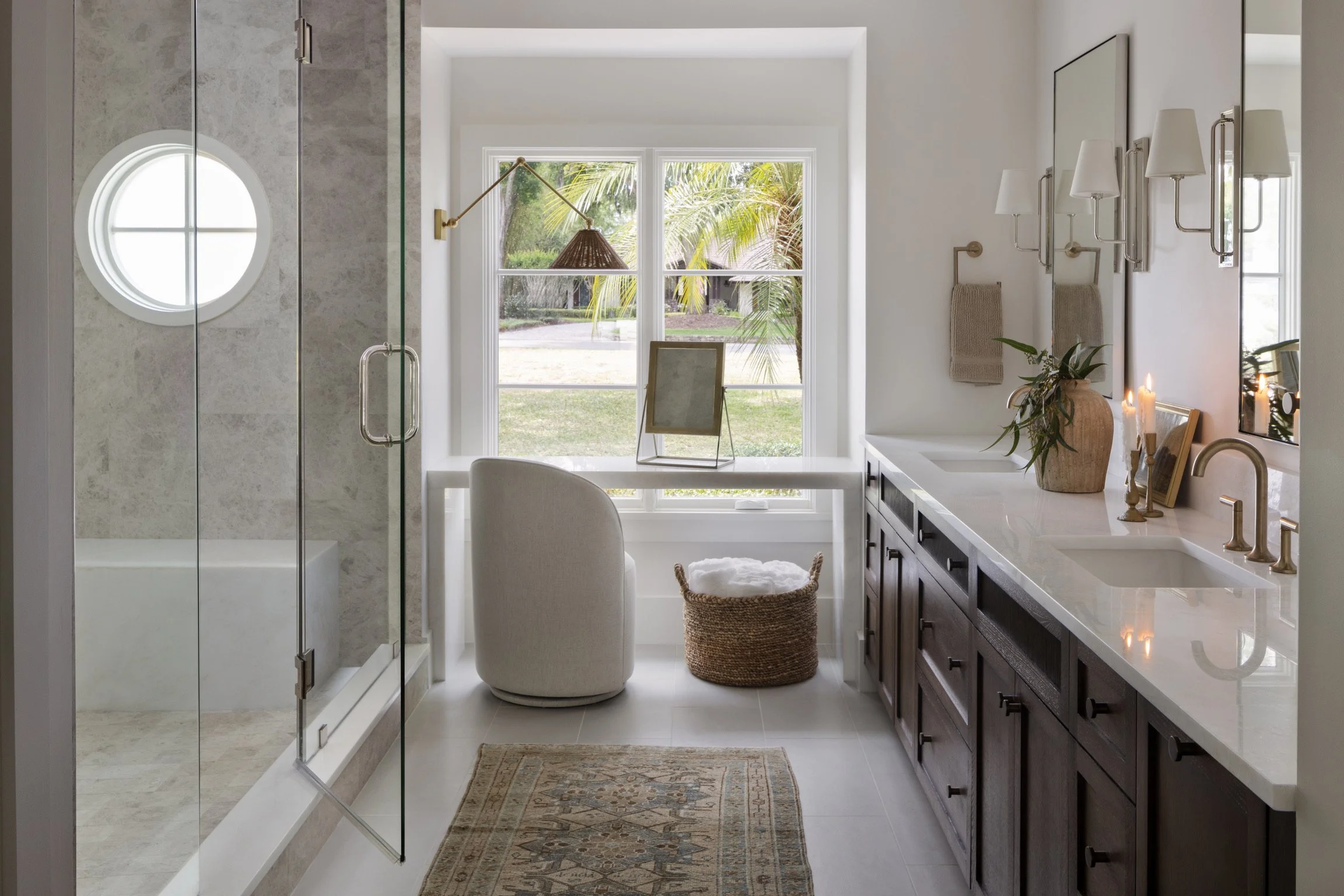 Modern bathroom with a glass shower, a large window, a white armchair, a woven laundry basket, a dark wood vanity with white countertop, candles, a plant, and wall-mounted lamps.