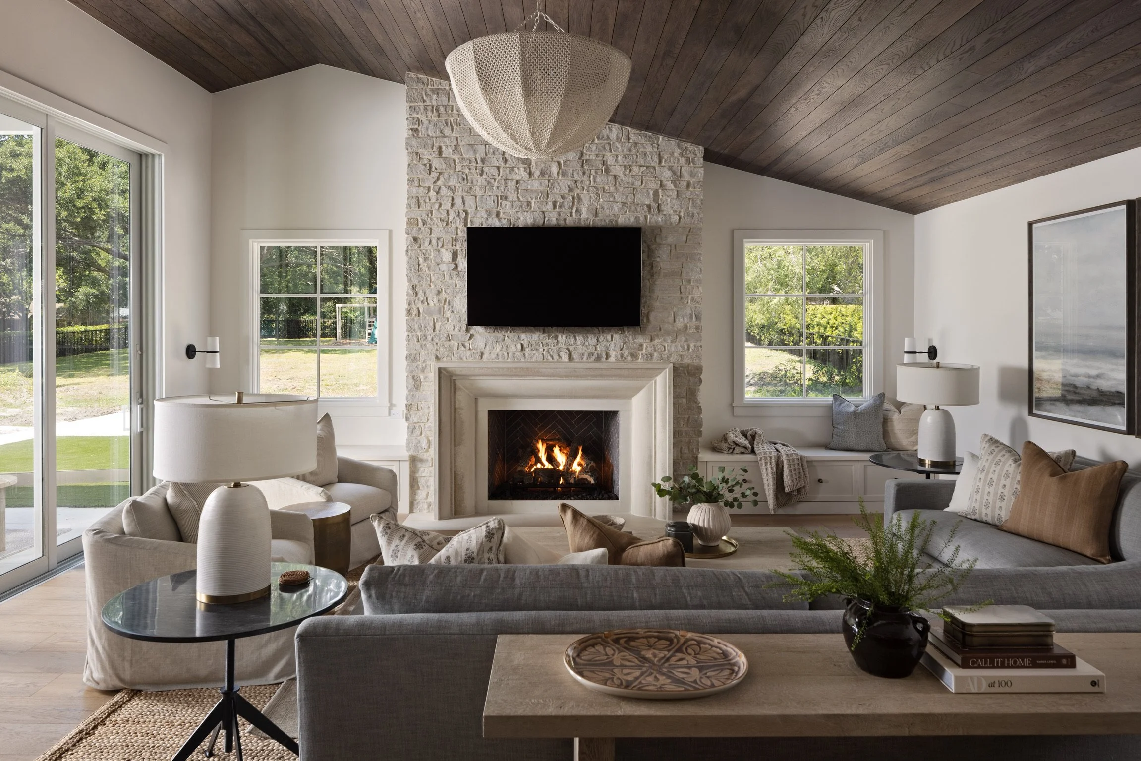 Living room with a wood-burning fireplace, a mounted TV above, and large windows showing a backyard view, decorated with neutral-colored furniture and decor.
