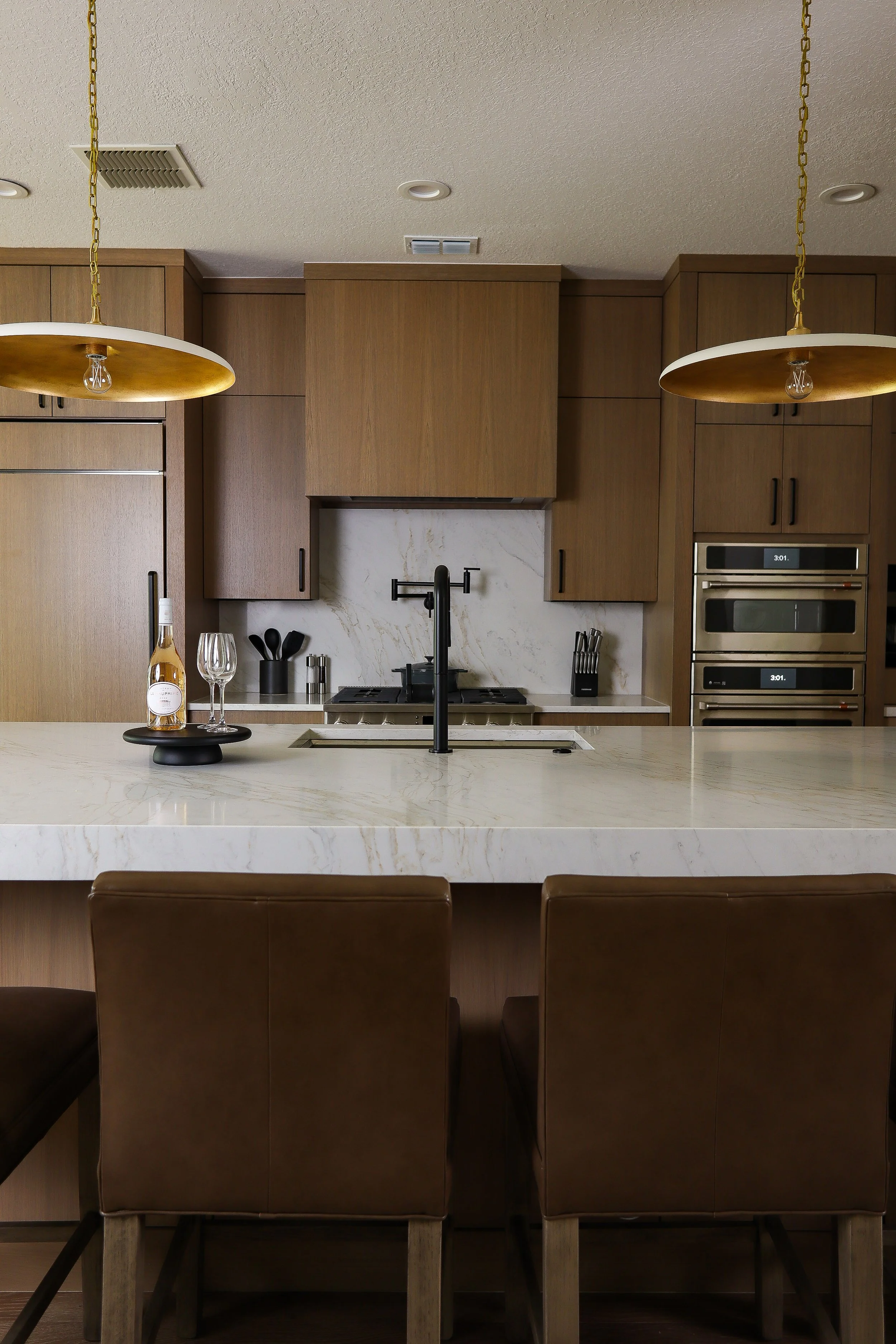 Modern kitchen with wooden cabinets, a white marble island, and two gold pendant lights. Behind the island, there is a stove, a marble backsplash, and built-in appliances. There is a tray with a bottle and two wine glasses on the island.