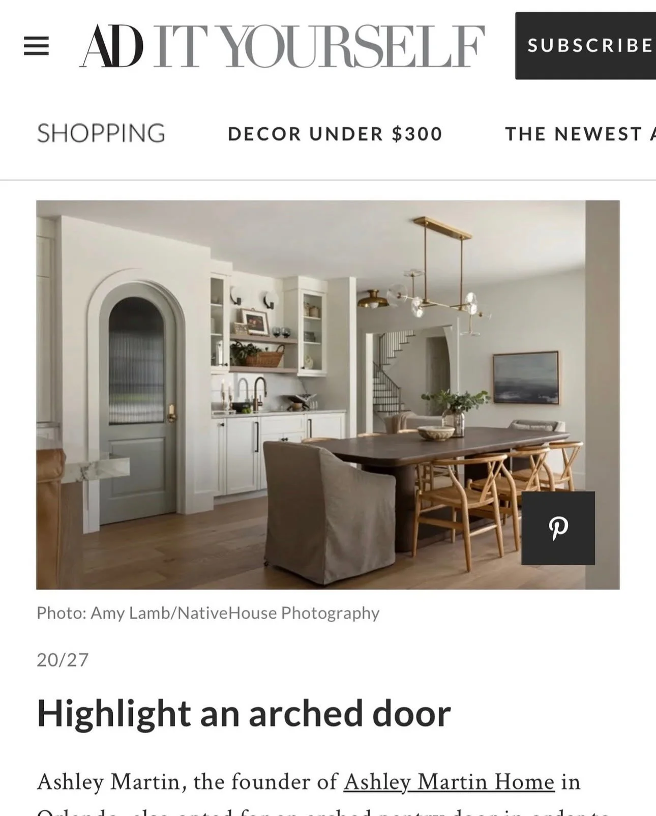 Interior view of a modern dining area featuring a large wooden dining table, chairs, a ceiling light fixture, a painting on the wall, and a staircase in the background.