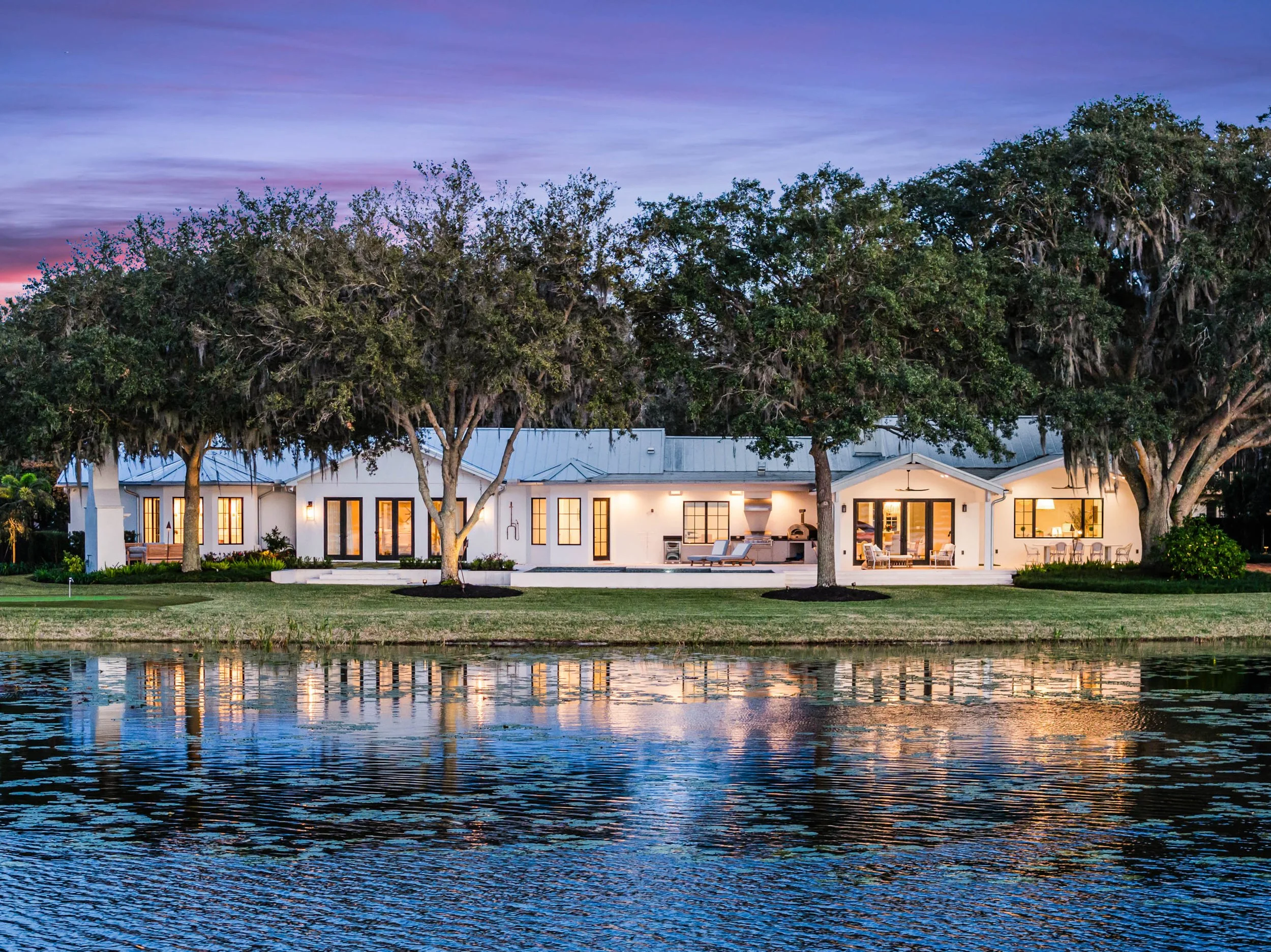 A modern white house with large windows and outdoor seating area, surrounded by trees, next to a body of water reflecting the house and trees during twilight.