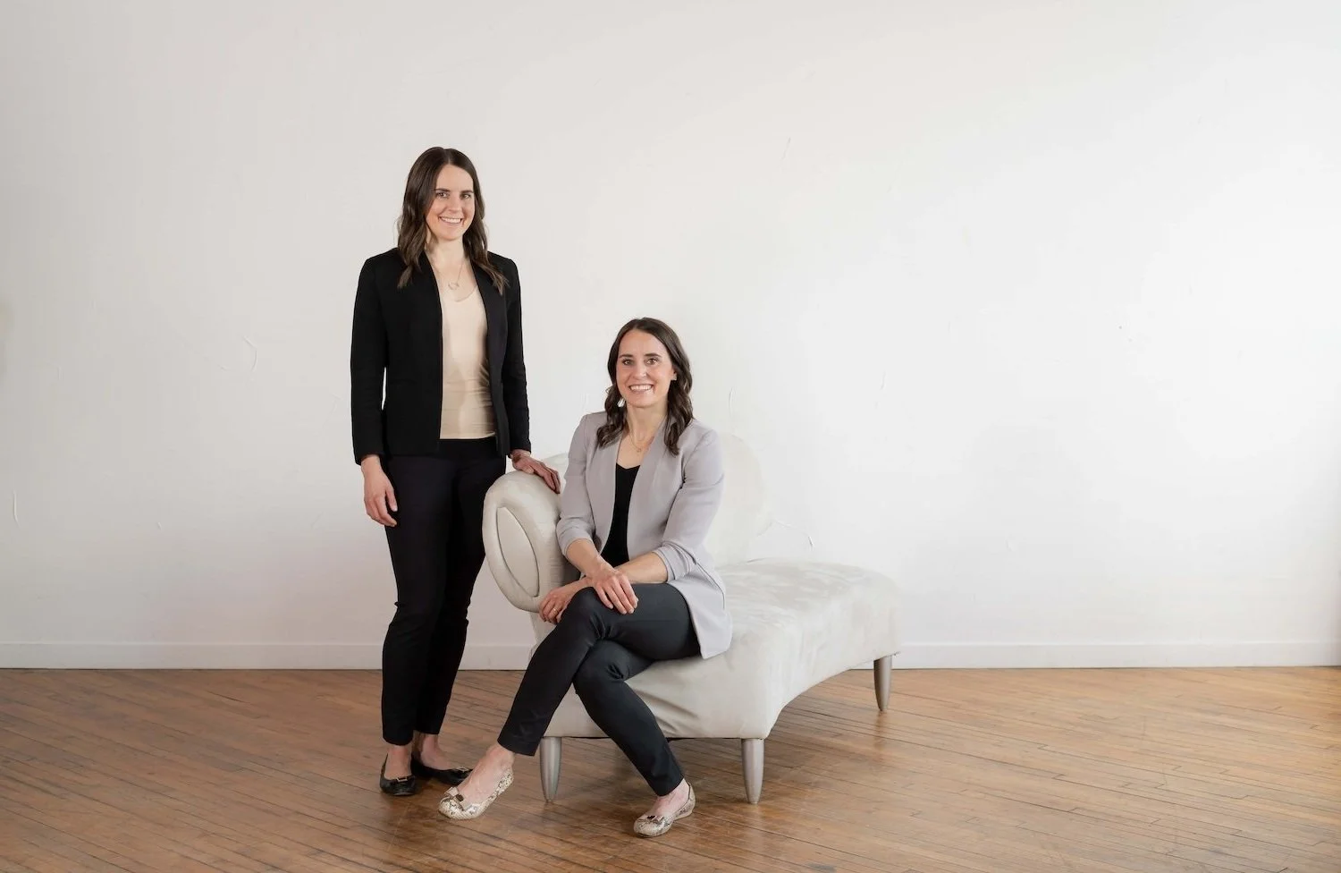 Two female doctors in Ada, MI in business attire, one standing, one sitting on a white couch, against a white wall and wooden floor.