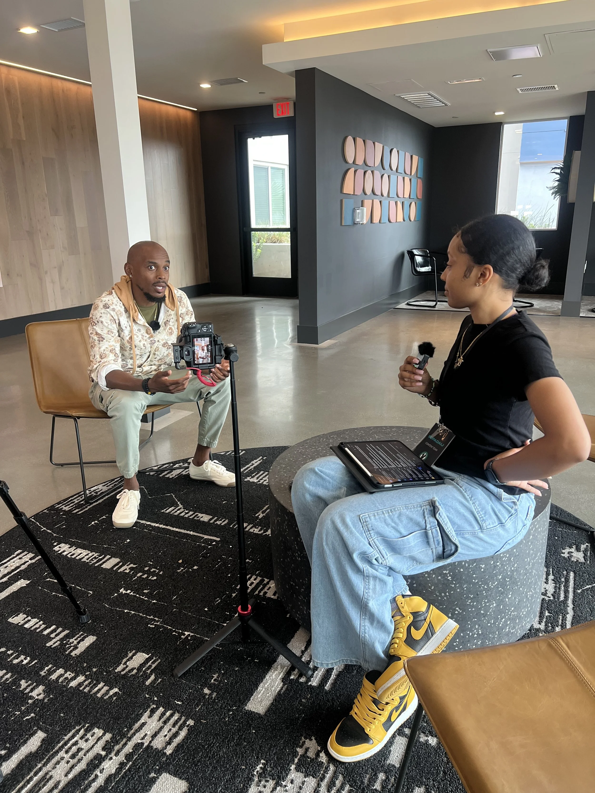 A man in a patterned shirt and light-colored pants is seated in a leather chair, talking to a woman who is standing and holding a microphone. The woman has dark hair tied back in a bun, and is wearing a black shirt, light denim jeans, and yellow sneakers. They are in a modern indoor space with black and wood-paneled walls, artwork, and large windows.