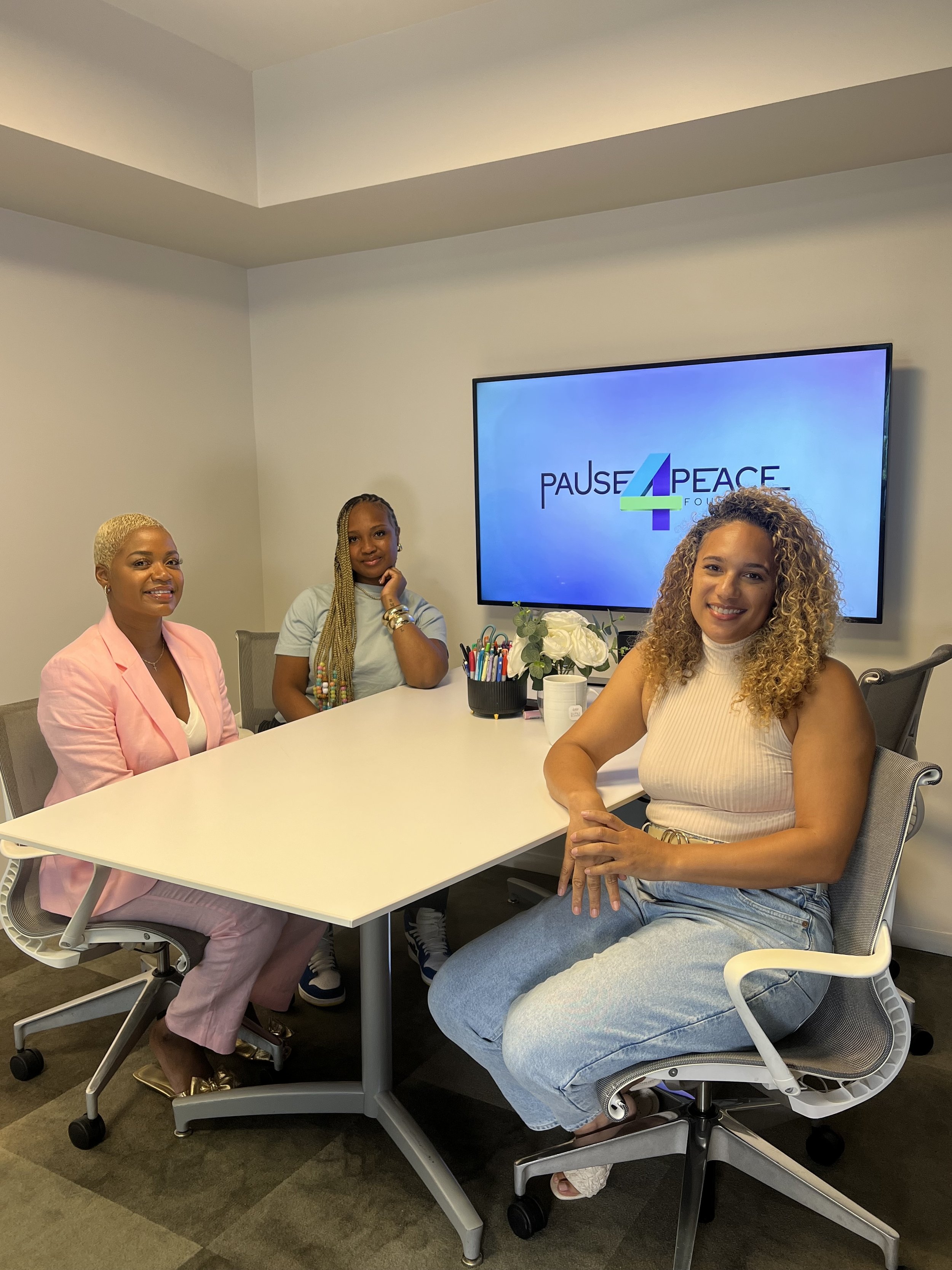 Four women sitting around a conference table in a meeting room. There is a large monitor on the wall displaying the 'Pause4Peace Foundation' logo, and a set of pens and flowers on the table.