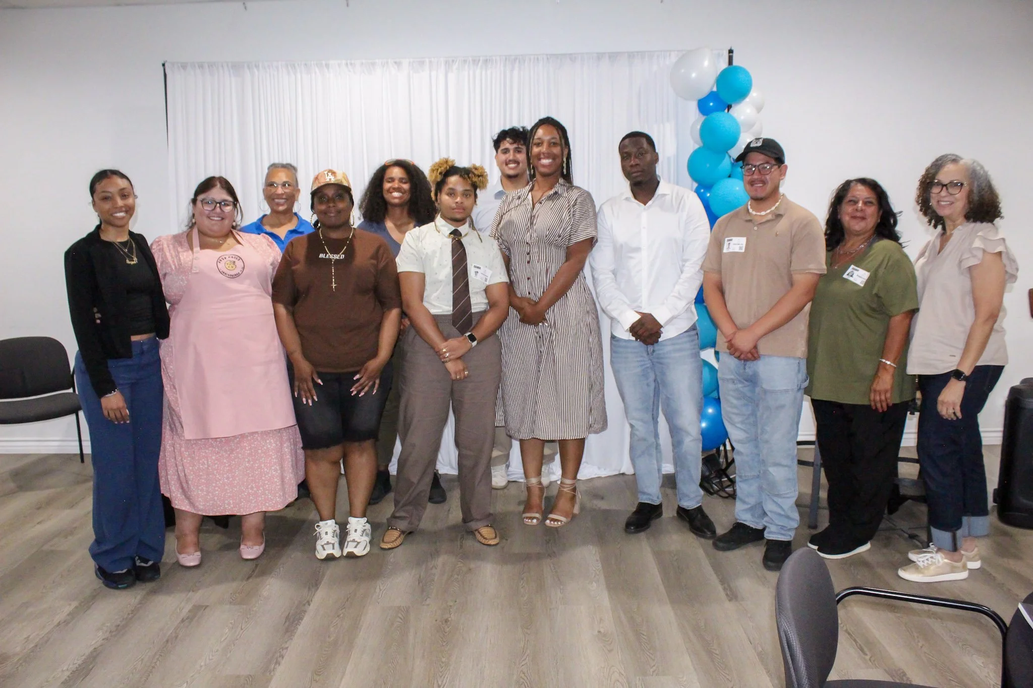 Group of diverse people standing together in an indoor setting with a white curtain and balloons in the background.