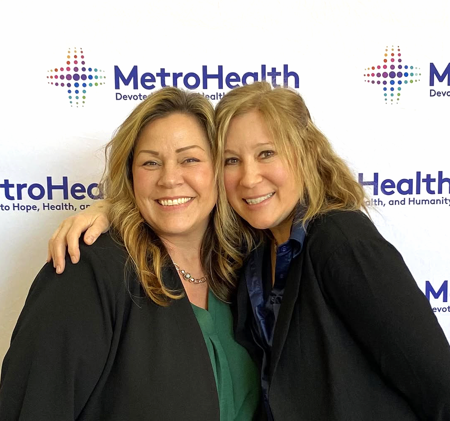 Cori Woolf and colleague embracing at MetroHealth event, standing in front of MetroHealth Devoted to Health branded step-and-repeat banner