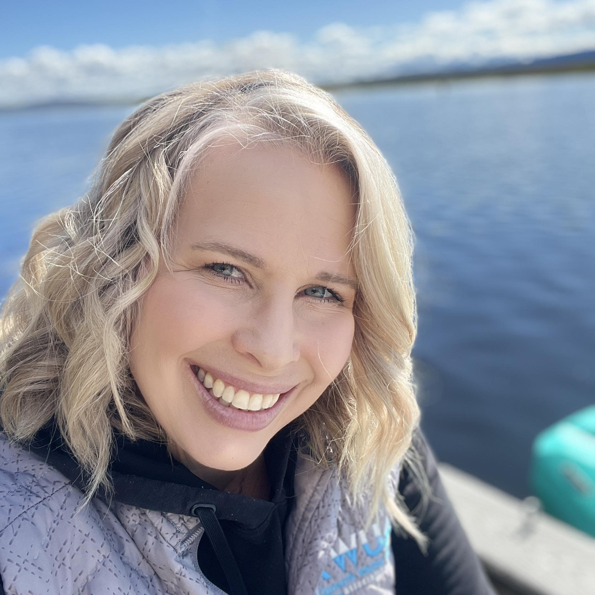 A woman with blonde hair and blue eyes smiling near a body of water with a clear sky.