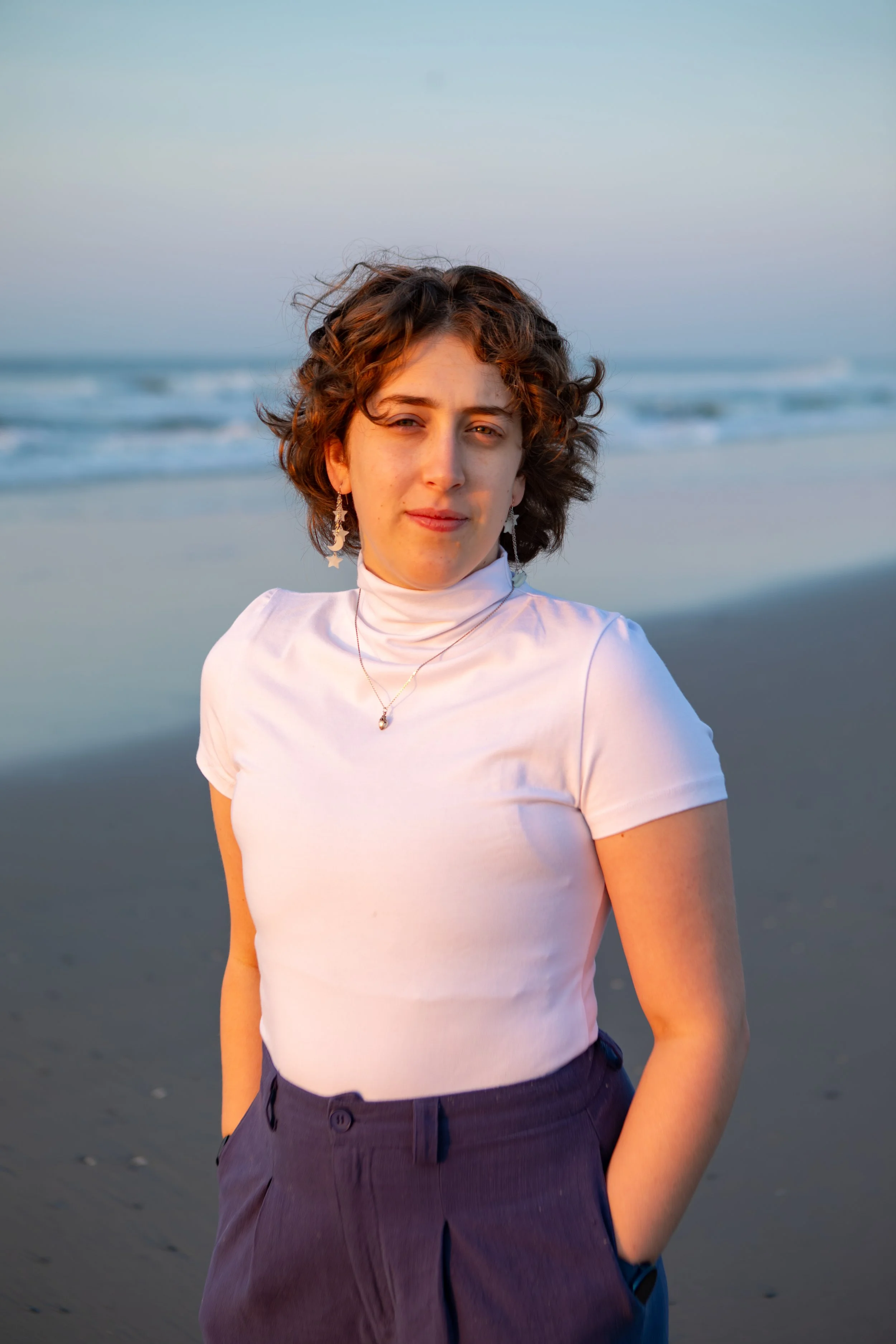 Jean stands on a beach at sunrise. They are a white person with curly, shoulder length brown hair. He wears a white turtleneck with short sleeves, dangly earrings, and a necklace.