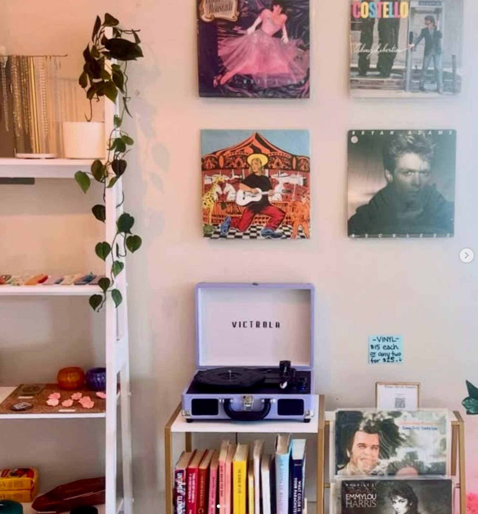 A wall with four colorful art posters, a white bookshelf with books and decorative items, a lavender record player on a small wooden table, and a smaller shelf with books and vinyl records.