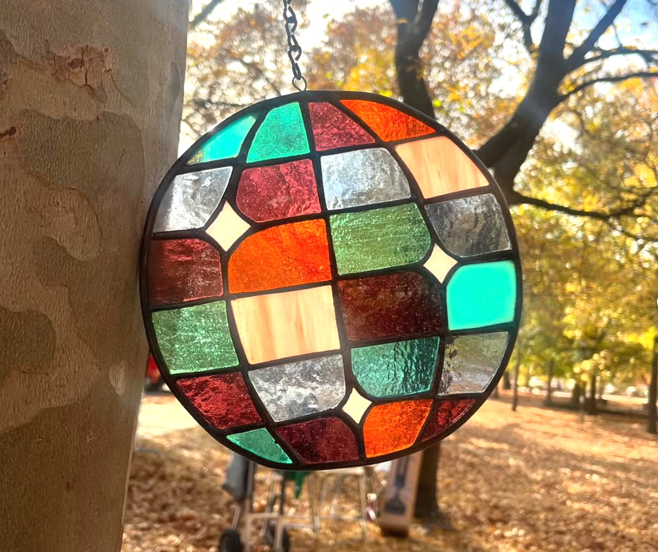 A round stained glass ornament hanging from a chain, with colorful geometric pieces, outdoors in a park during autumn with fallen leaves and trees with yellow and orange foliage.