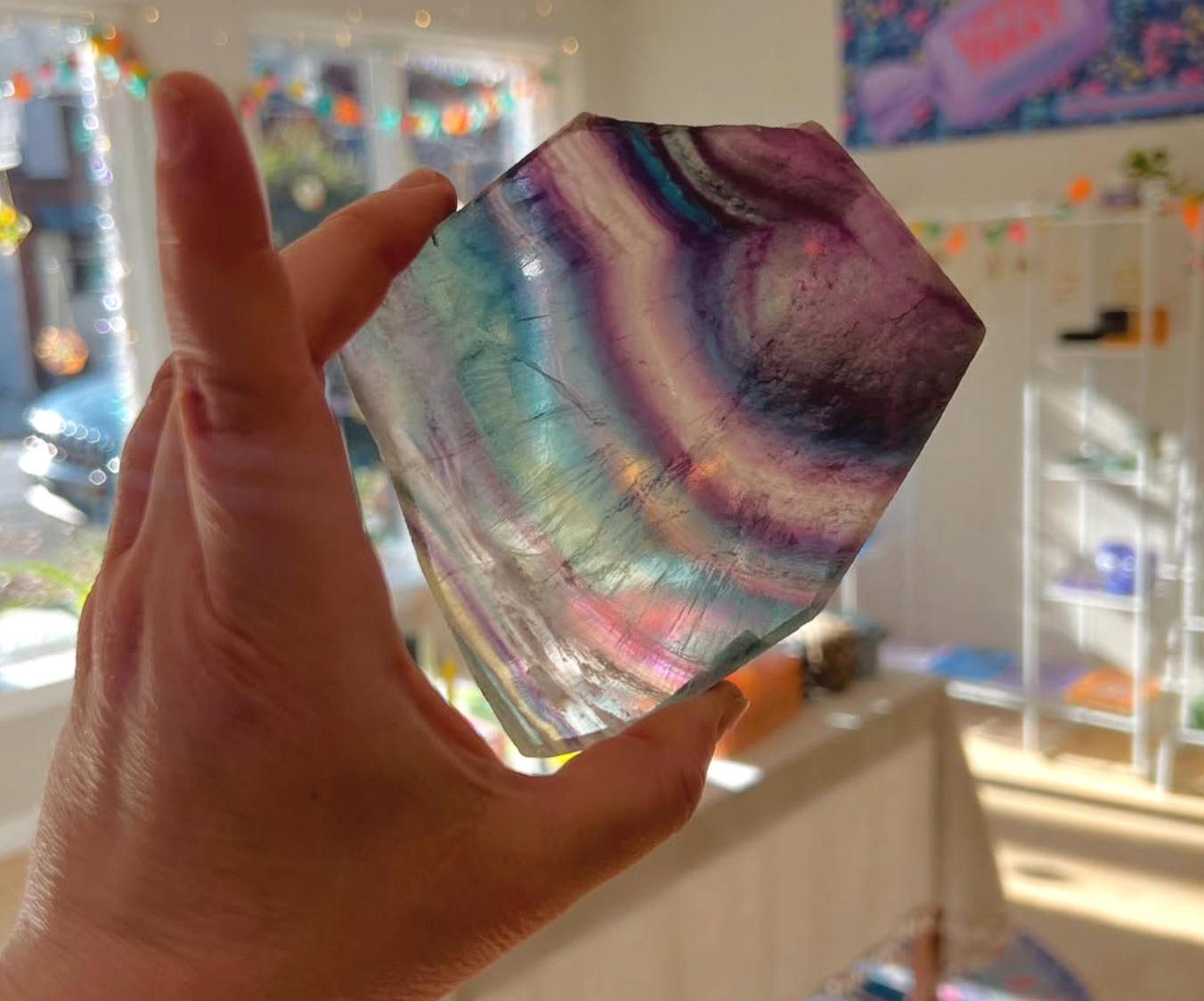 A hand holding a large, iridescent, multi-colored piece of obsidian with swirled purple, green, and blue patterns inside a brightly lit store with shelves and colorful decorations in the background.