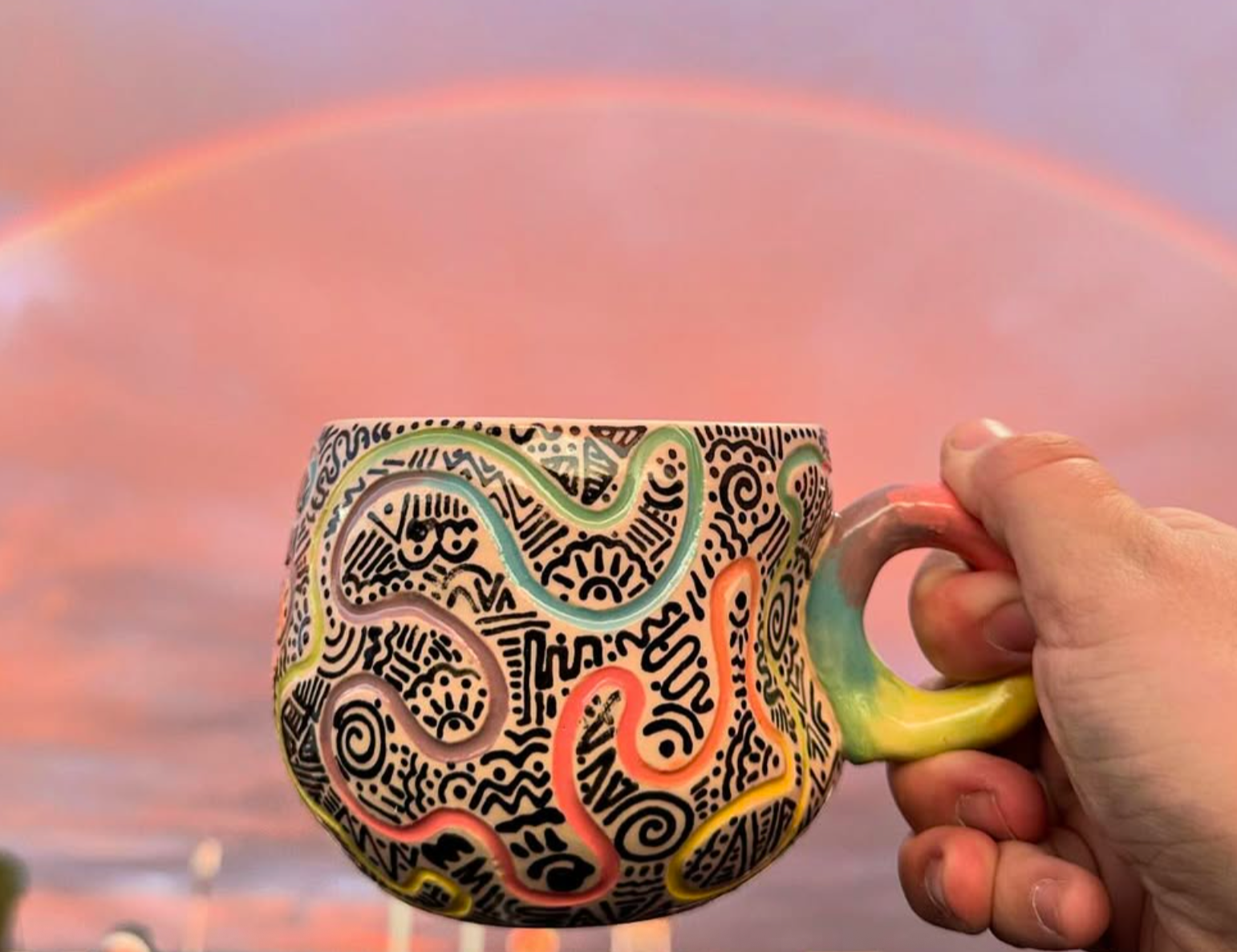 A colorful, abstract-patterned ceramic mug held by a person's hand against a soft pink and purple sky with a faint rainbow.