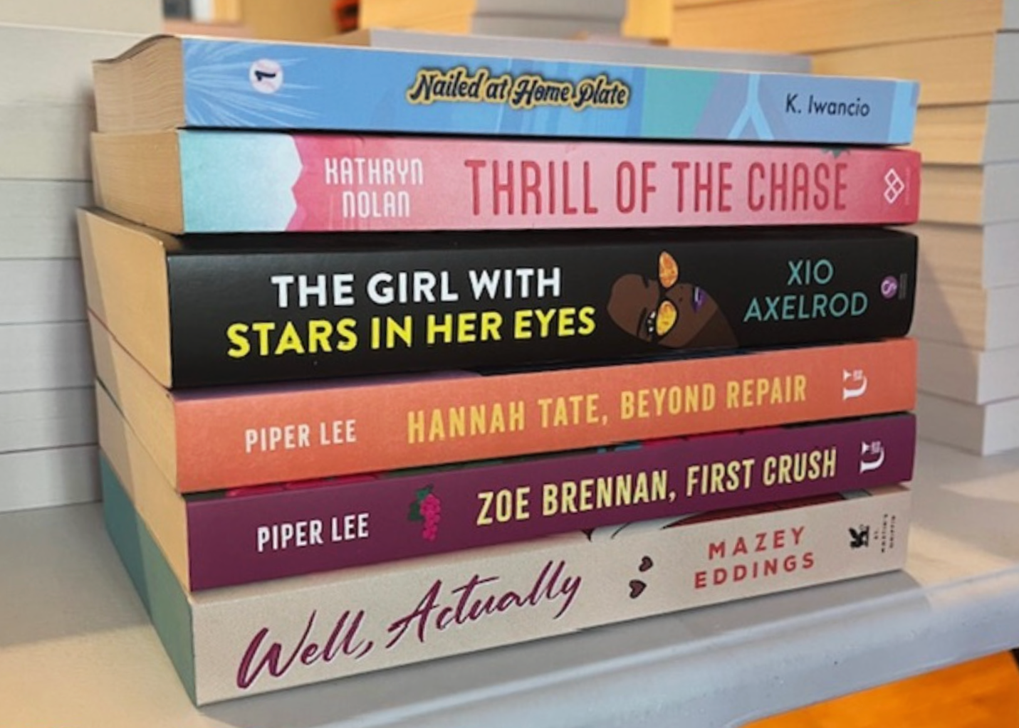 Stack of six books with colorful covers leaning against a white wooden wall. The titles include "Nailed at Home Plate" by K. Iwancio, "Thrill of the Chase" by Kathryan Nolan, "The Girl with Stars in Her Eyes" by Xio Axelrod, "Hannah Tate, Beyond Repair" by Piper Lee, "Zoe Brennan, First Crush" by Piper Lee, and "Well, Actually" by Mazey Eddings.