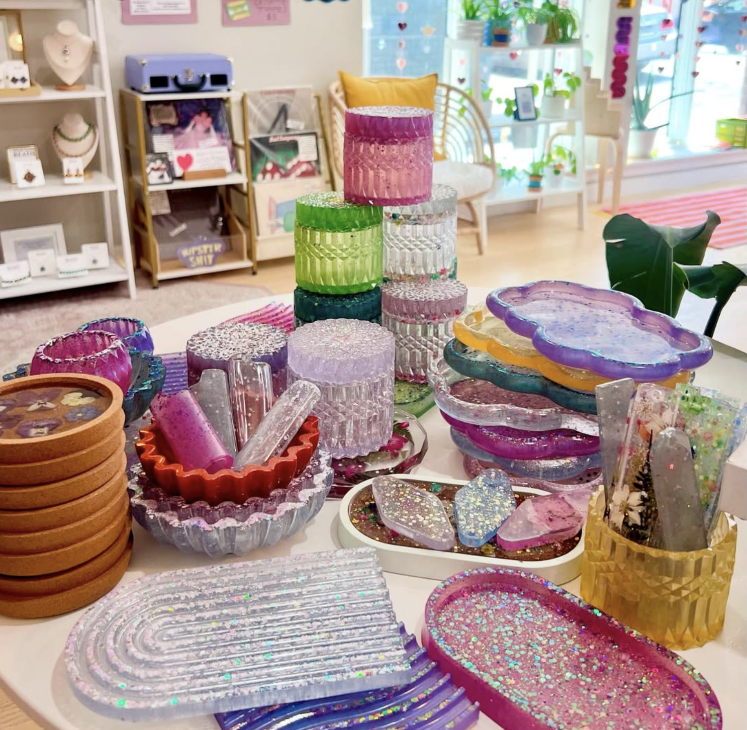 Colorful glass and resin jewelry display on a table in a boutique shop, with shelves and windows decorated with plants in the background.