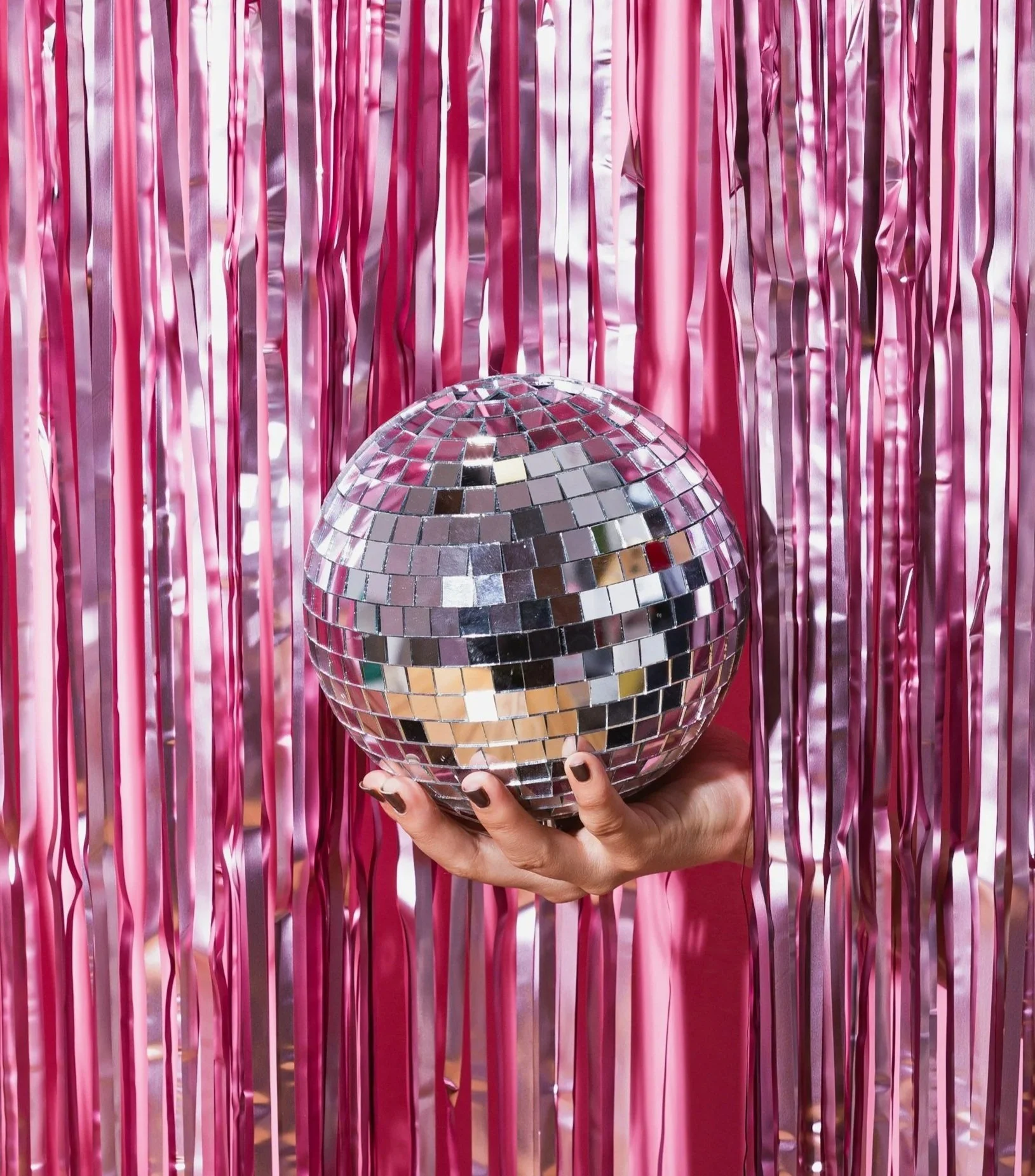 A person holding a disco ball in front of pink metallic streamers background.