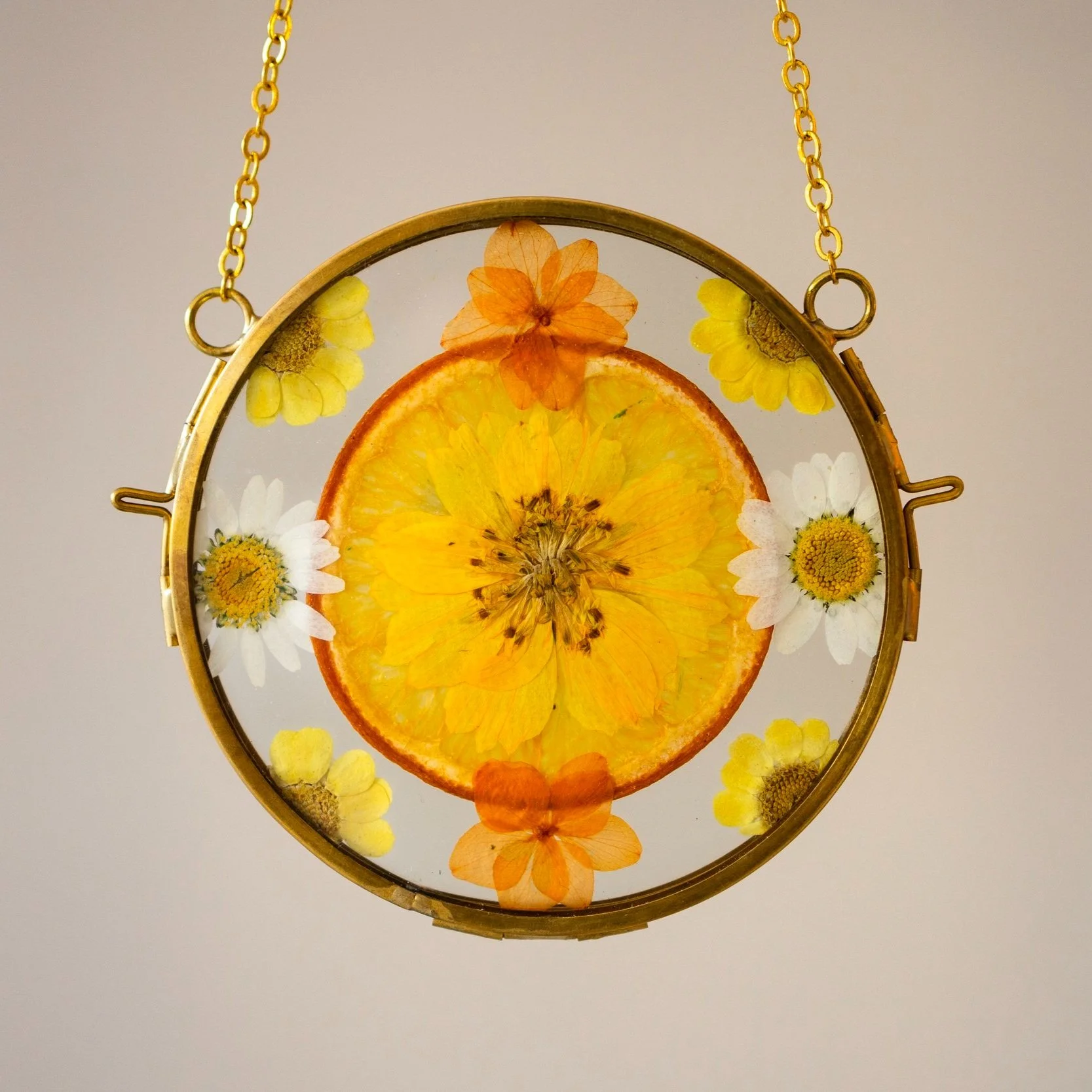 Gold pendant necklace with a circular frame filled with a dried orange slice, surrounded by various pressed flowers including white daisies, orange and yellow flowers, and a small green flower, on a plain background.