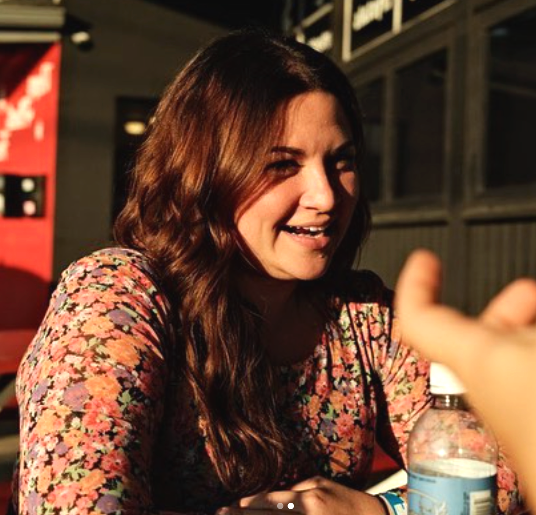 A woman with long, wavy brown hair smiling and talking to someone off-camera. She is wearing a floral-patterned top and sitting at a table with a water bottle in front of her. The background suggests a cozy restaurant or cafe setting.