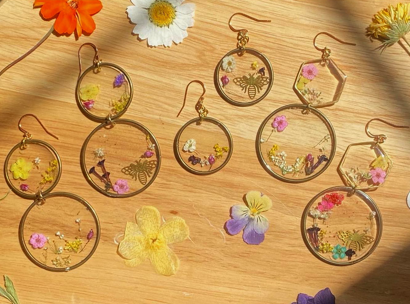 Set of six round and one hexagonal earrings with gold outlines, filled with pressed flowers and bee or butterfly charms, arranged on a wooden surface with scattered real flowers.
