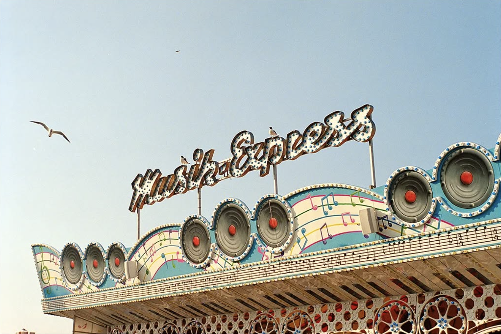 A vintage amusement park ride with a sign reading 'Music Express', colorful musical notes, and large red speakers, with seagulls flying in the sky.