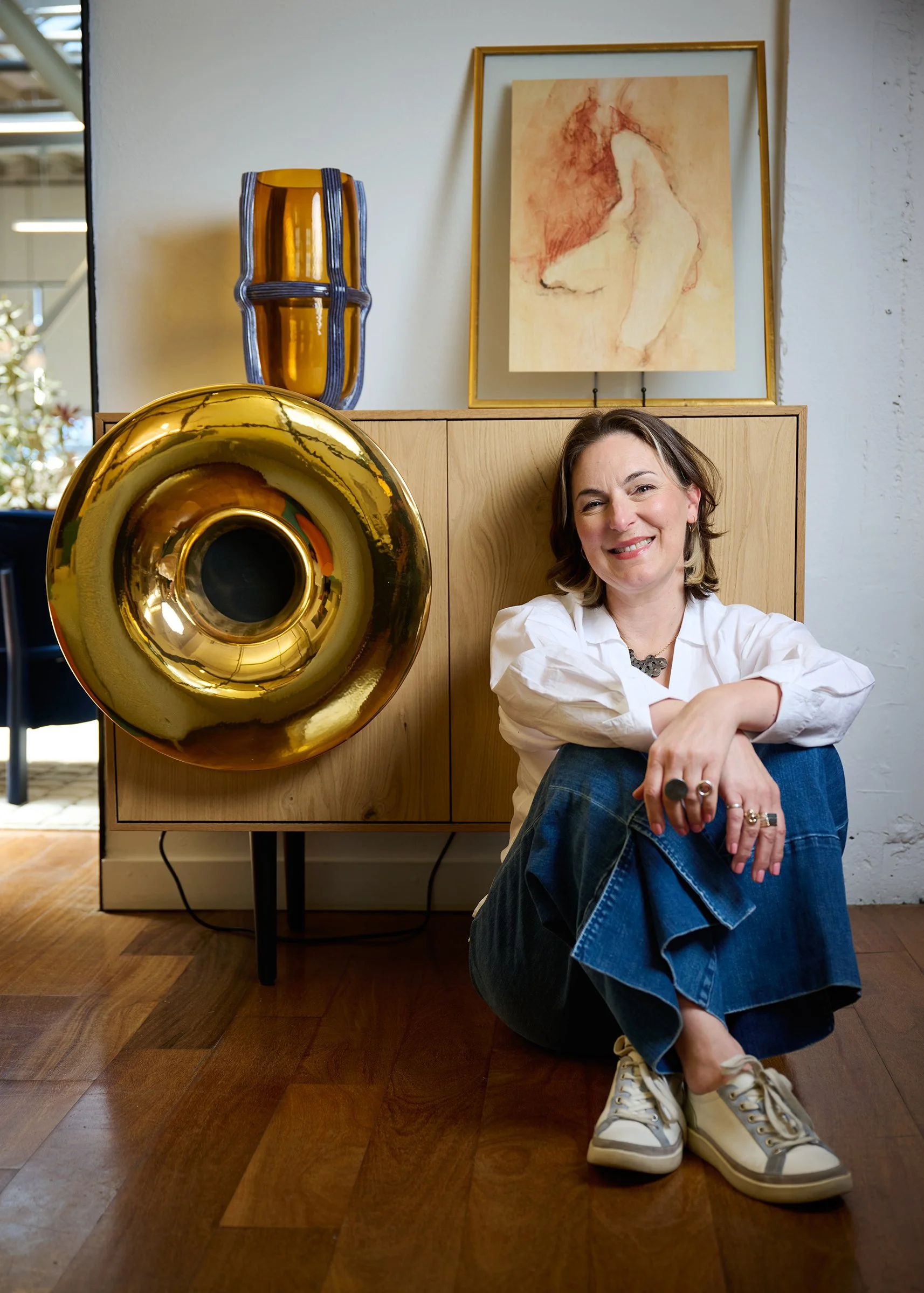 Smiling woman seated on a wood floor beside a cabinet displaying sculptural glass, brass, and framed artwork.