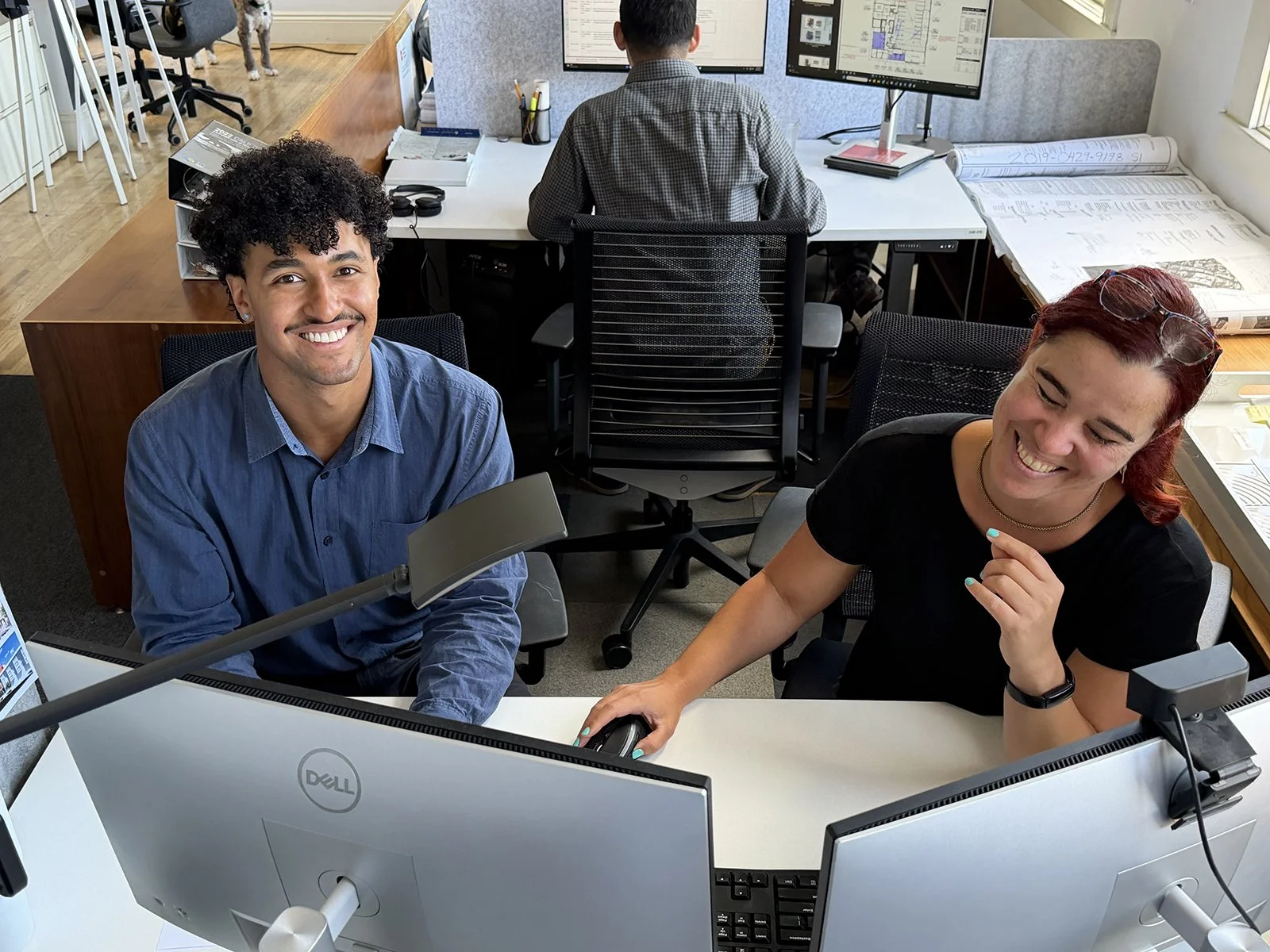 Two Y.A. studio team members smiling at a shared workstation in the office, with another colleague working at dual monitors in the background