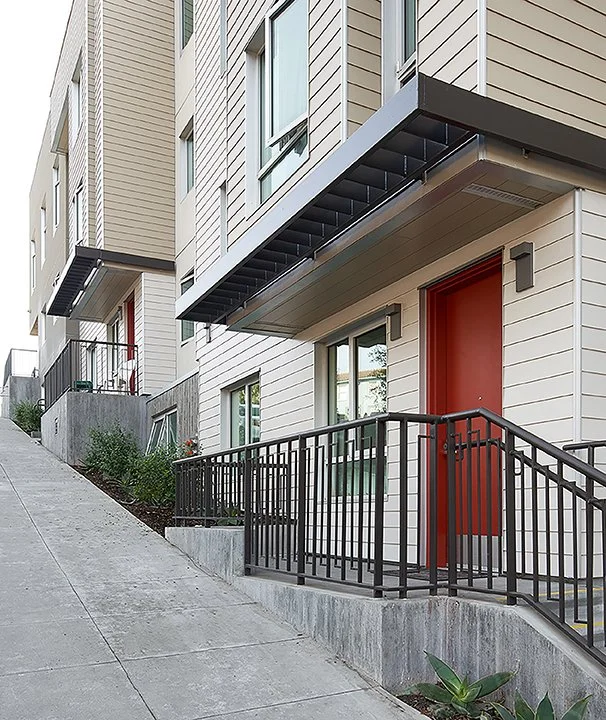 Accessible residential entrances with red doors, metal canopies, and railings along a steep sidewalk.