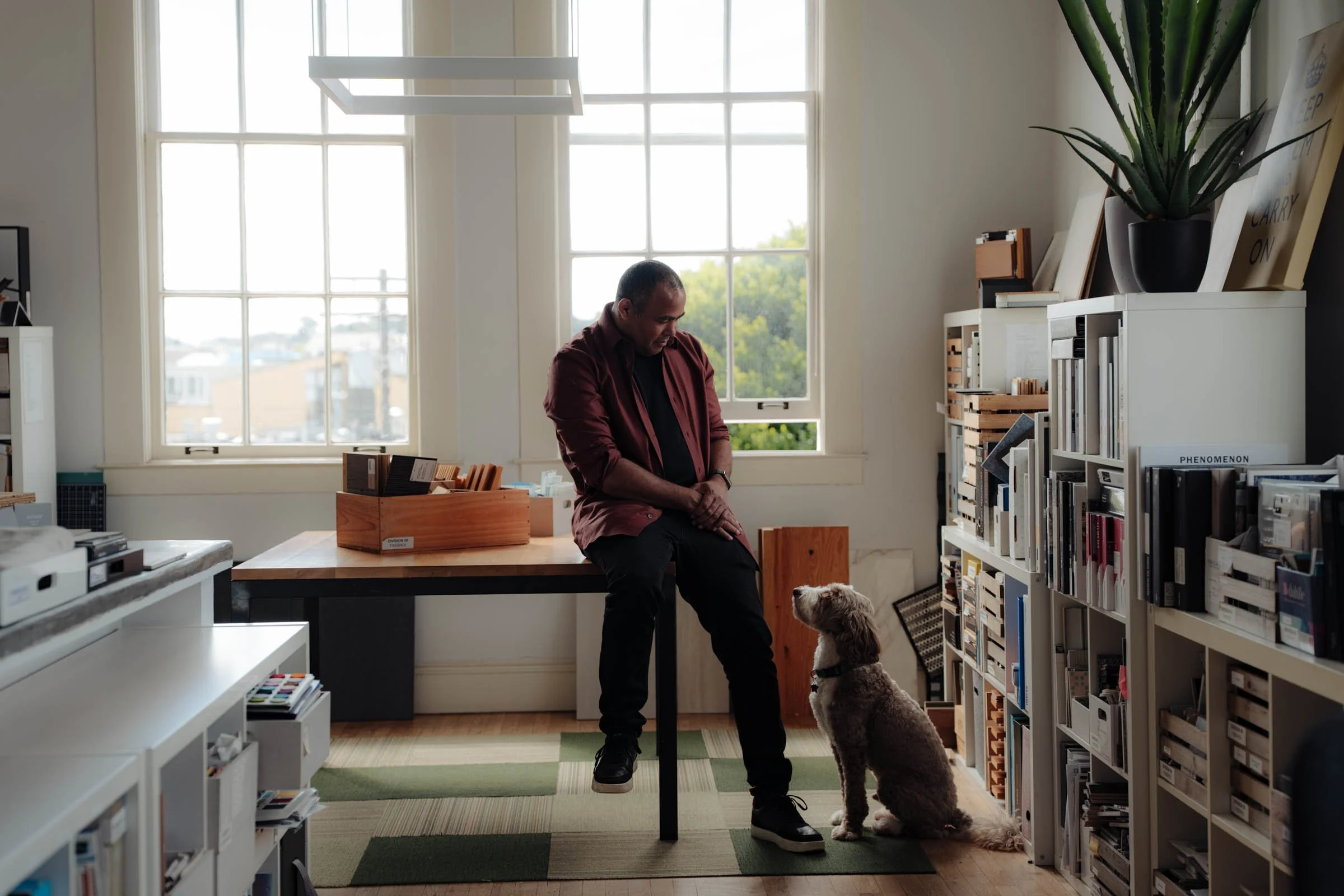 Man seated on a worktable looks down at a dog in a bright design studio lined with material samples, books, and storage shelves