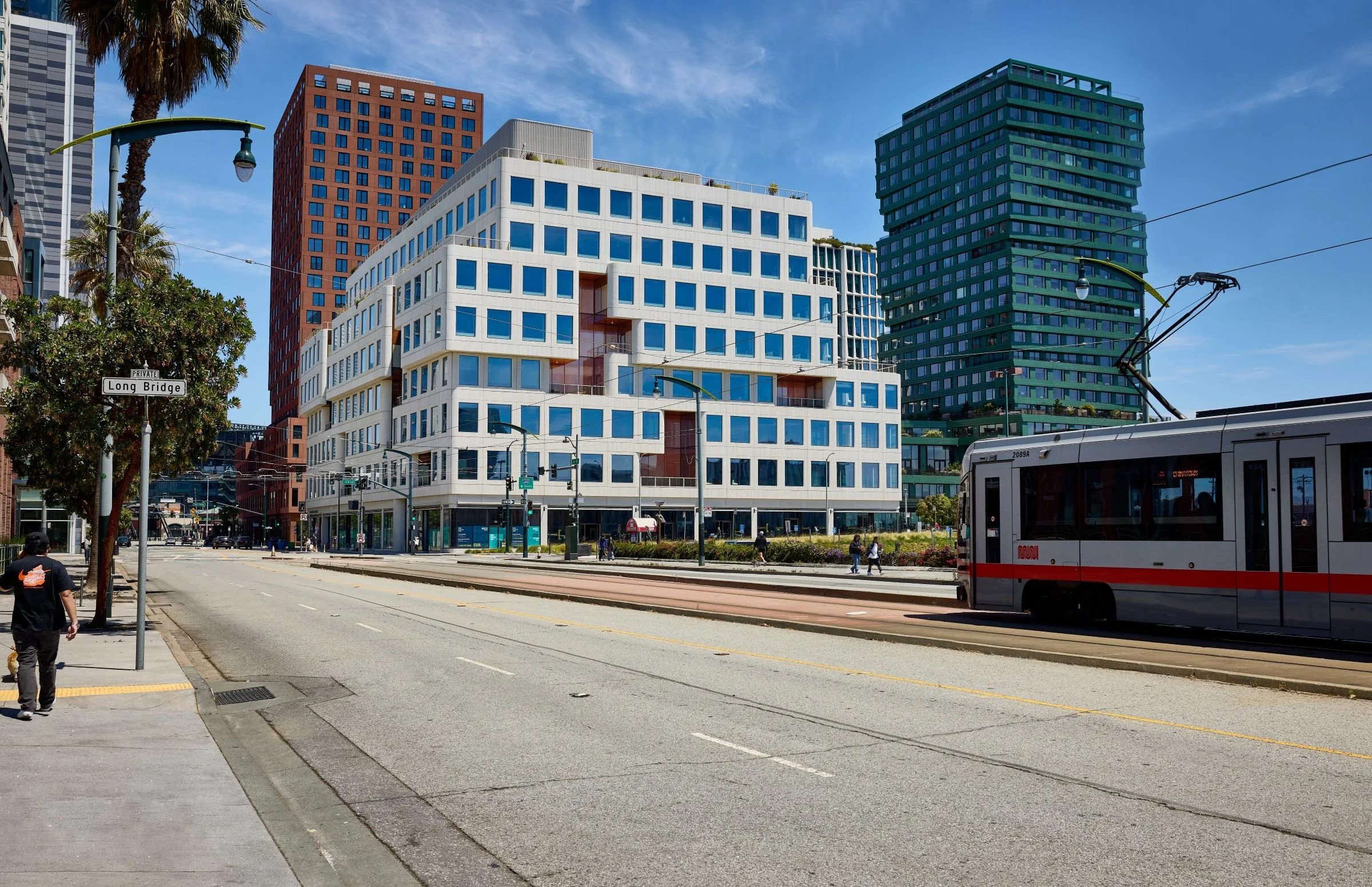 Mission Rock streetscape with stepped white building, residential towers, and Muni light rail along Third Street.